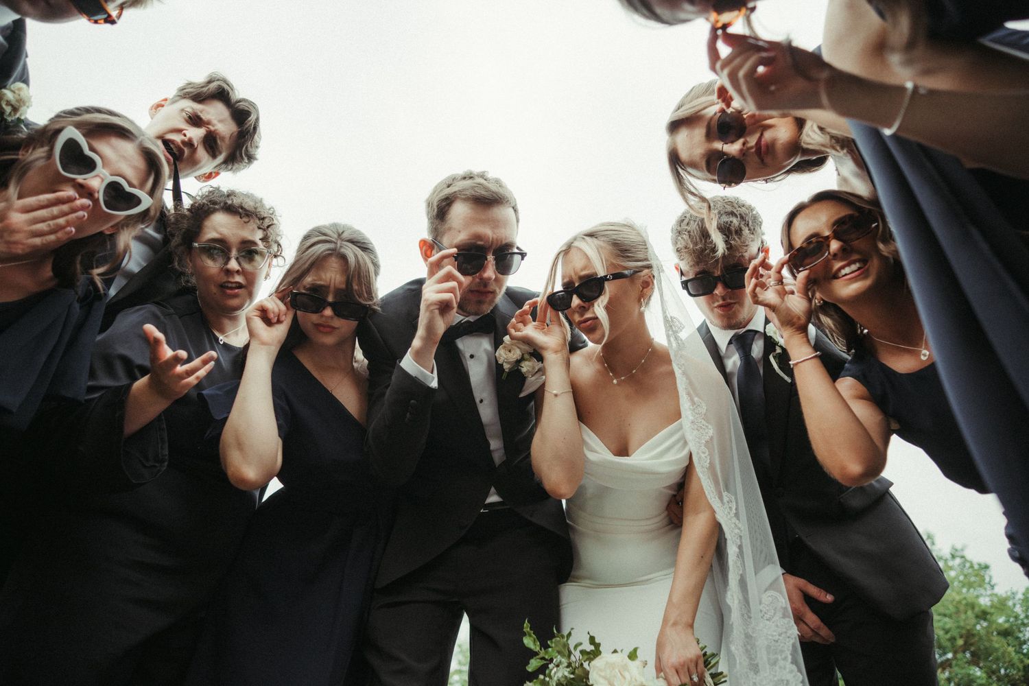 Bride, groom, and wedding party posing fun photo all wearing heart-shaped sunglasses outdoors at wedding celebration.