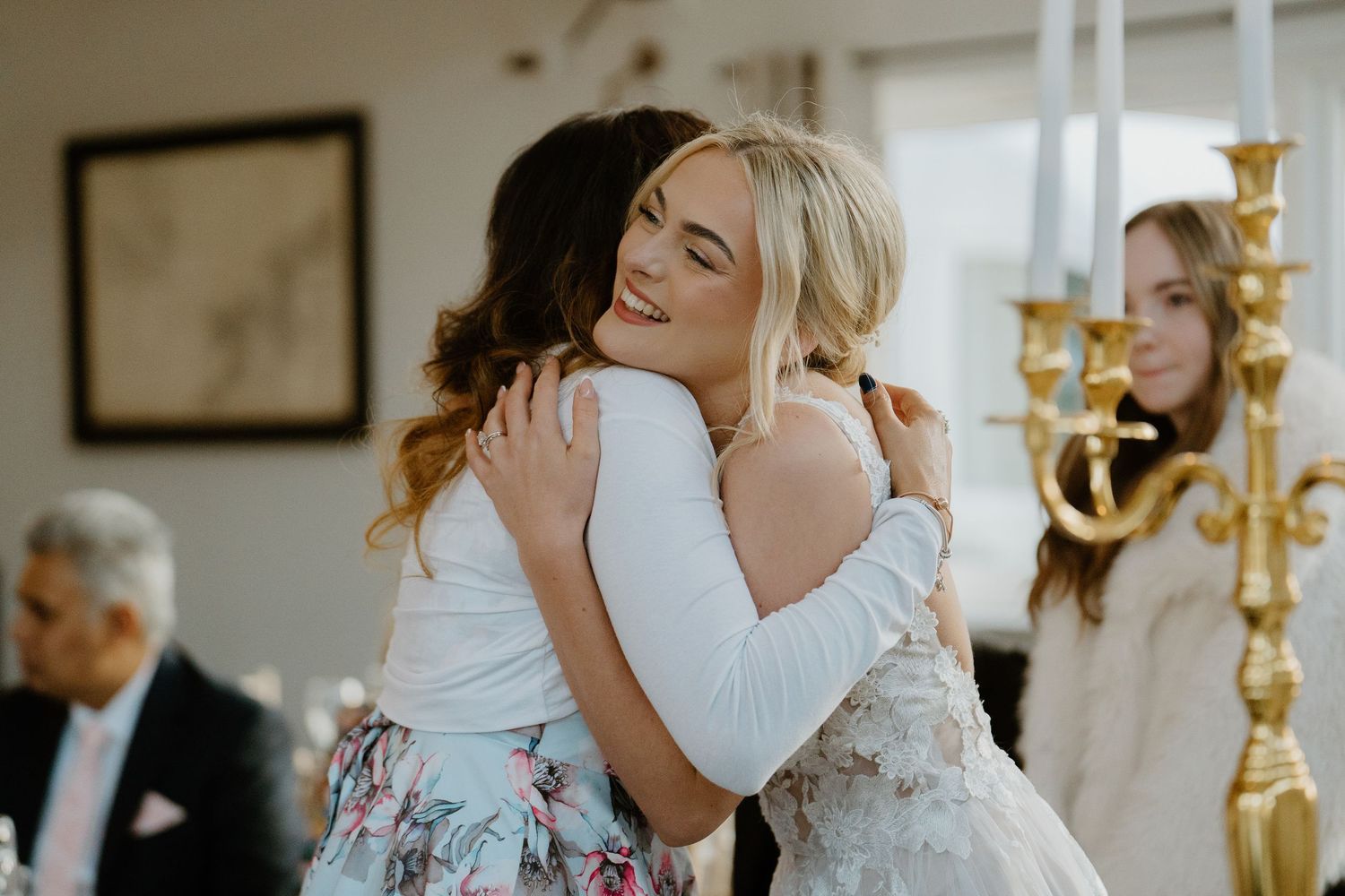 Two women in elegant attire sharing a warm embrace at a wedding reception with candles in background.