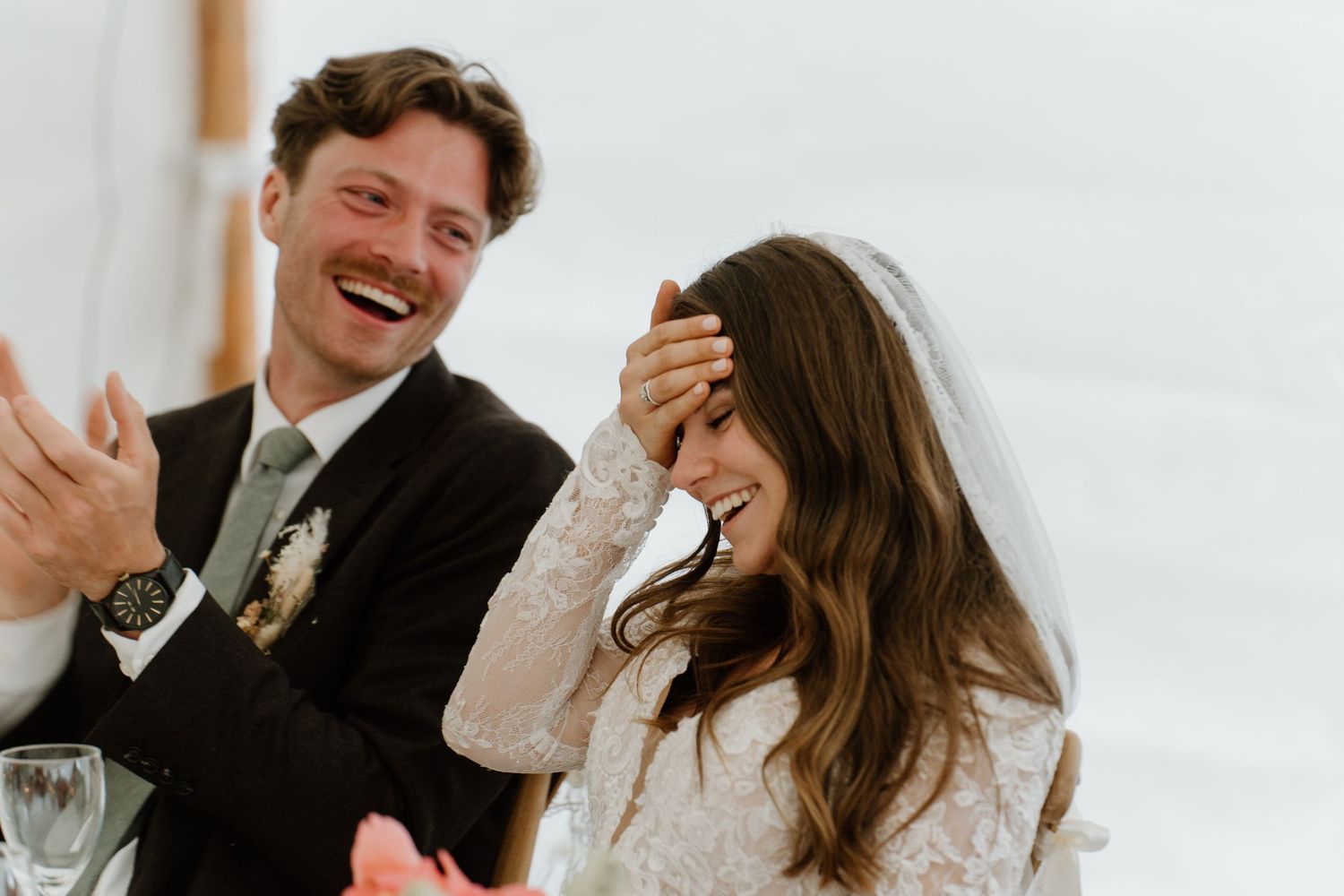 Bride covering her face laughing while groom smiles during wedding reception speeches.
