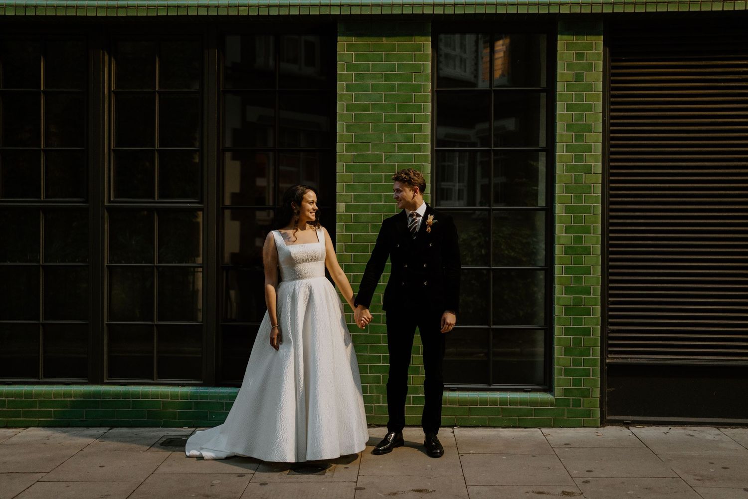 A bride and groom hold hands standing before a green tiled building facade on their wedding day.
