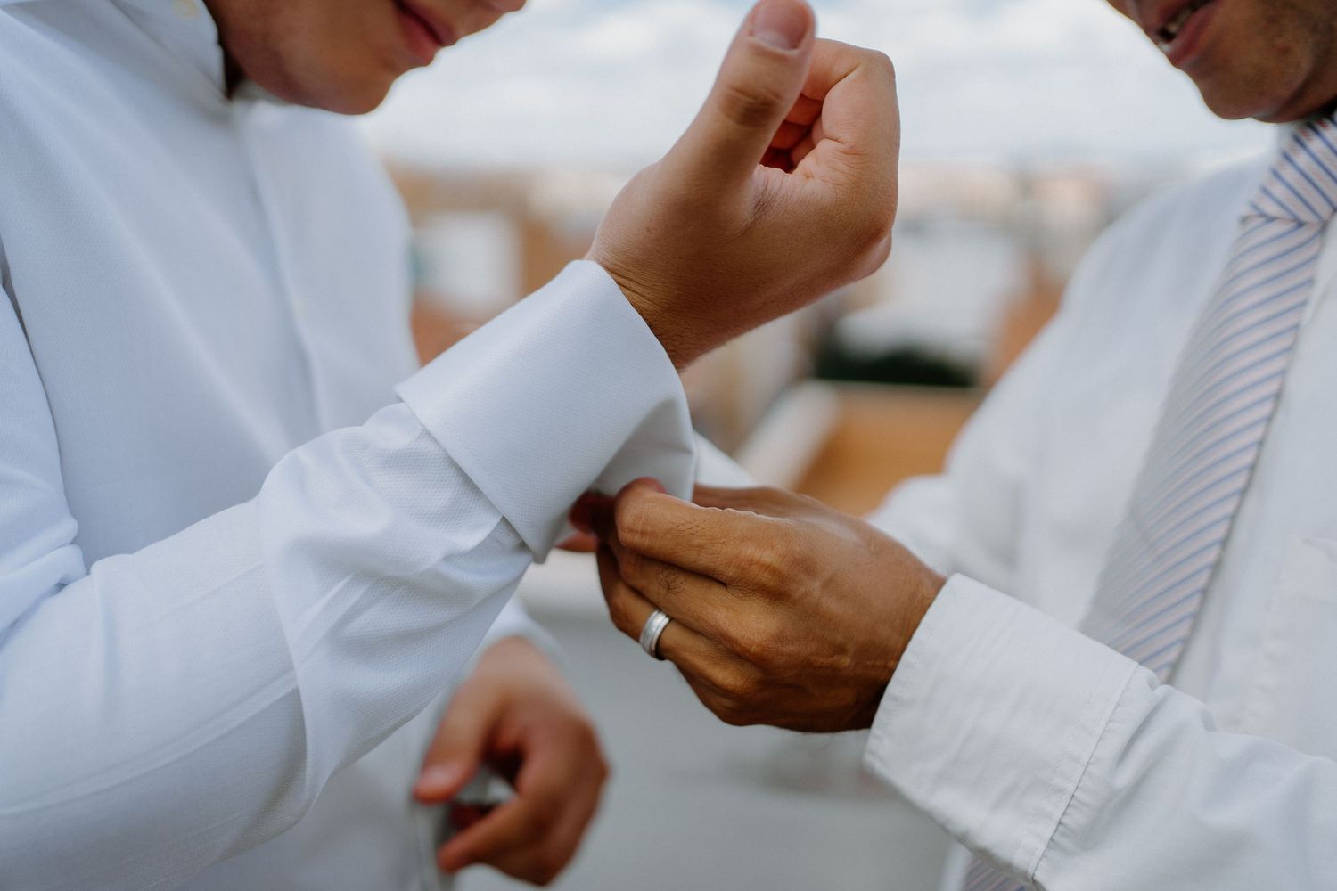 Groomsman helping groom fasten cufflinks on white dress shirt before wedding ceremony outdoors.