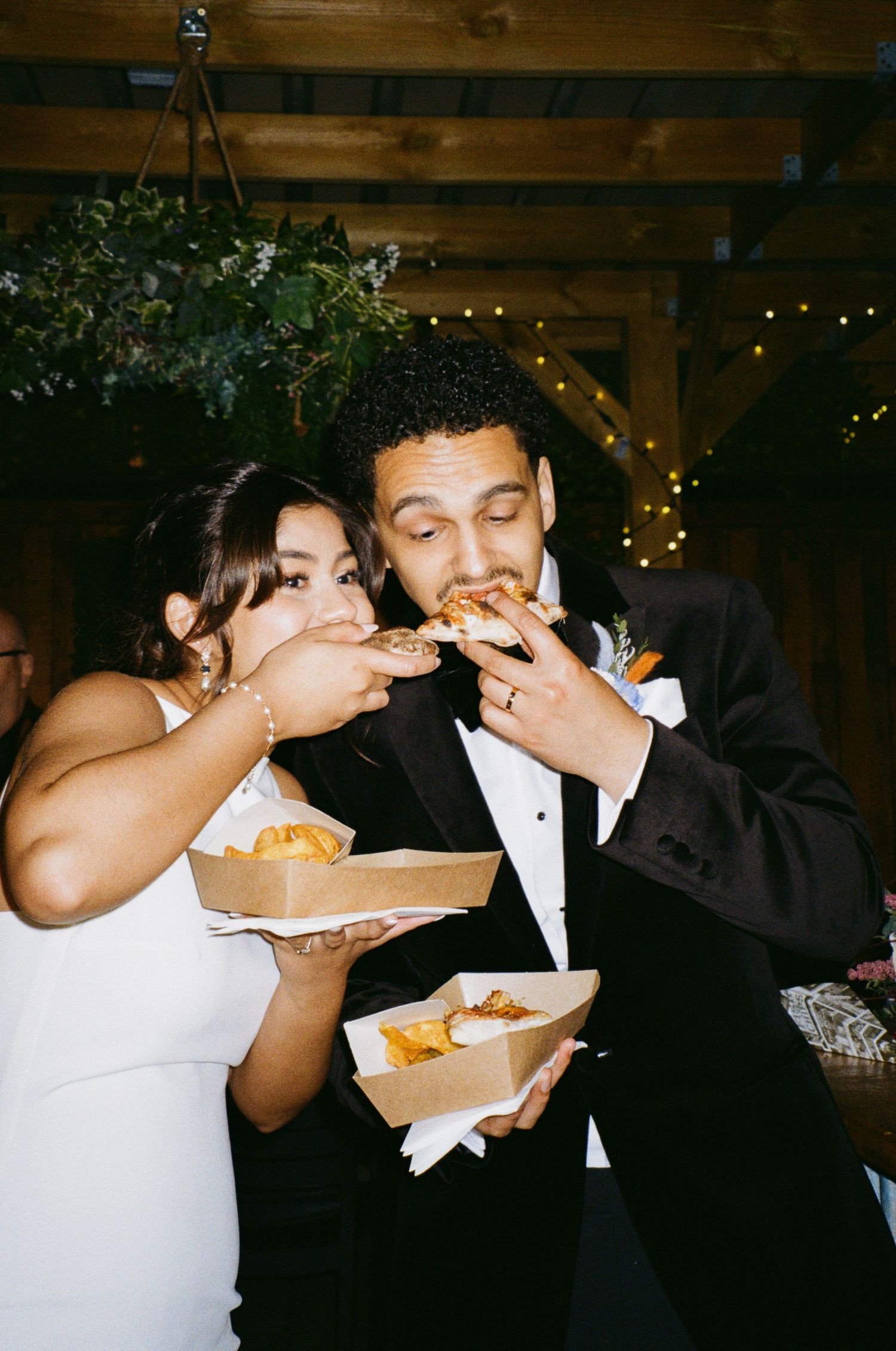 Bride and groom joyfully eating food together at their wedding reception under string lights.
