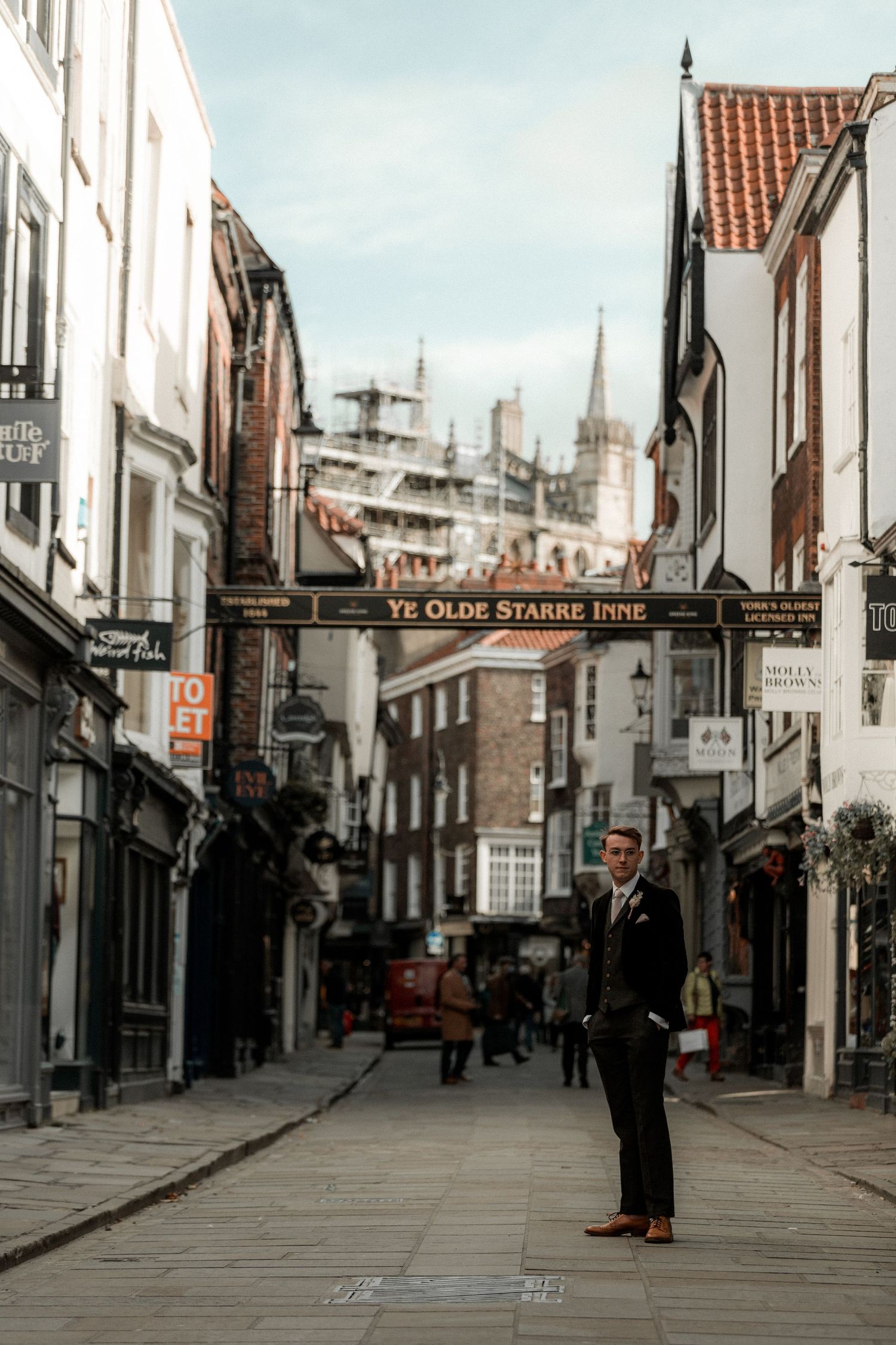 A historic cobblestone street in York with traditional shops and the cathedral spire visible in the distance.