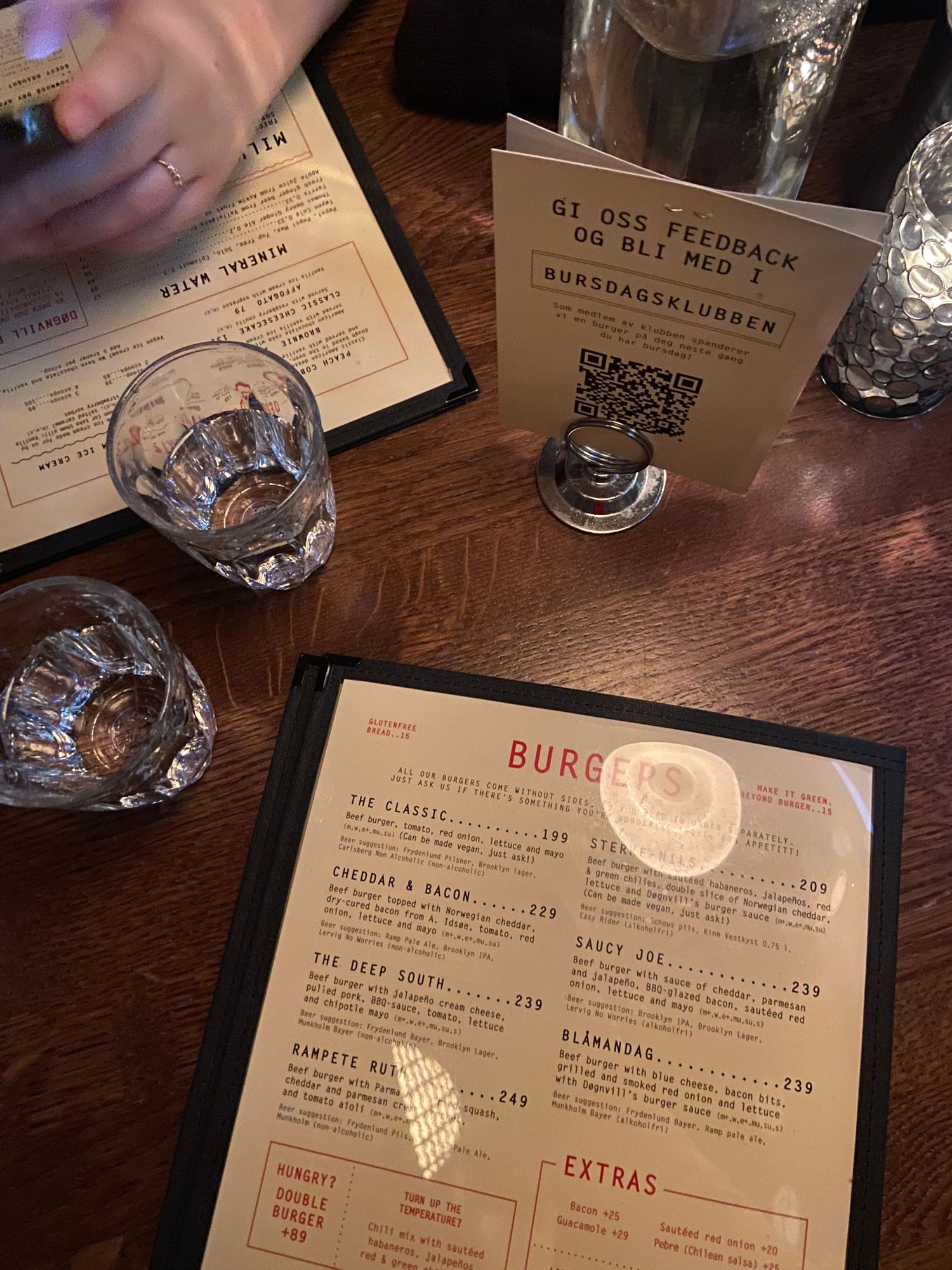 Restaurant table with menu showing burger options and QR code stand with empty cocktail glasses on wooden surface.