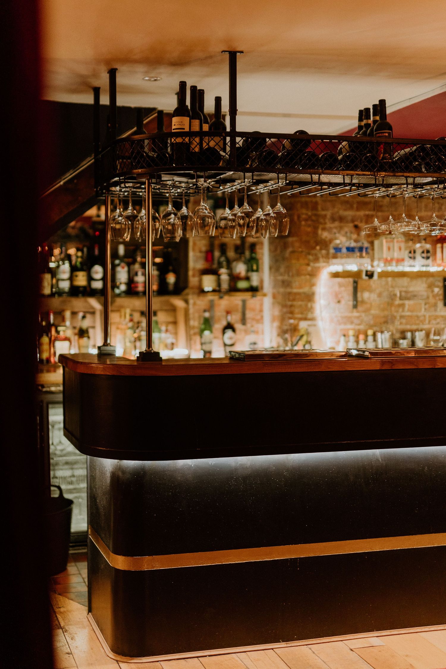 A warmly lit bar interior with hanging wine glasses, wooden counter, and shelves of bottles.