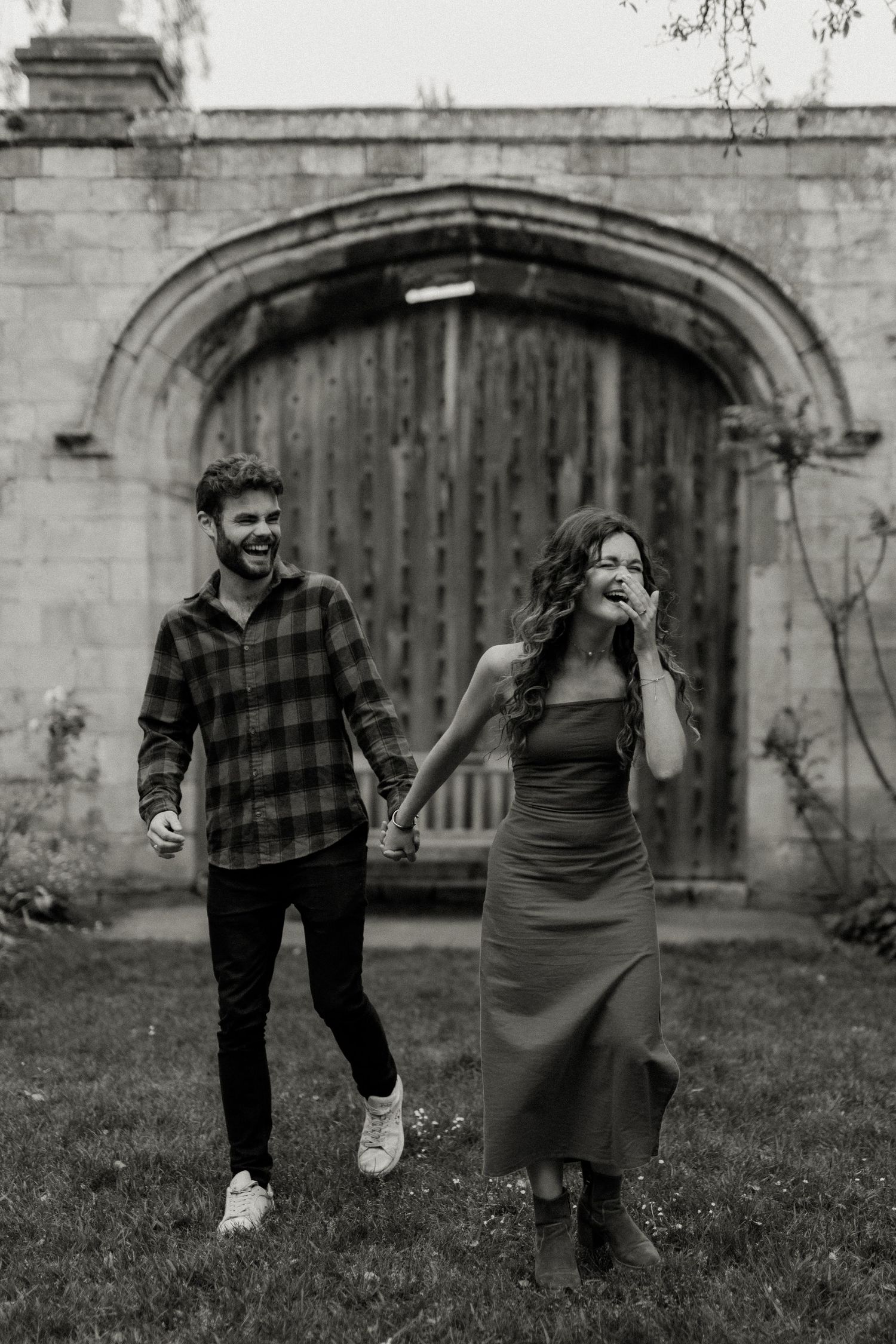 Black and white photo of a laughing couple holding hands while walking in front of a stone archway.