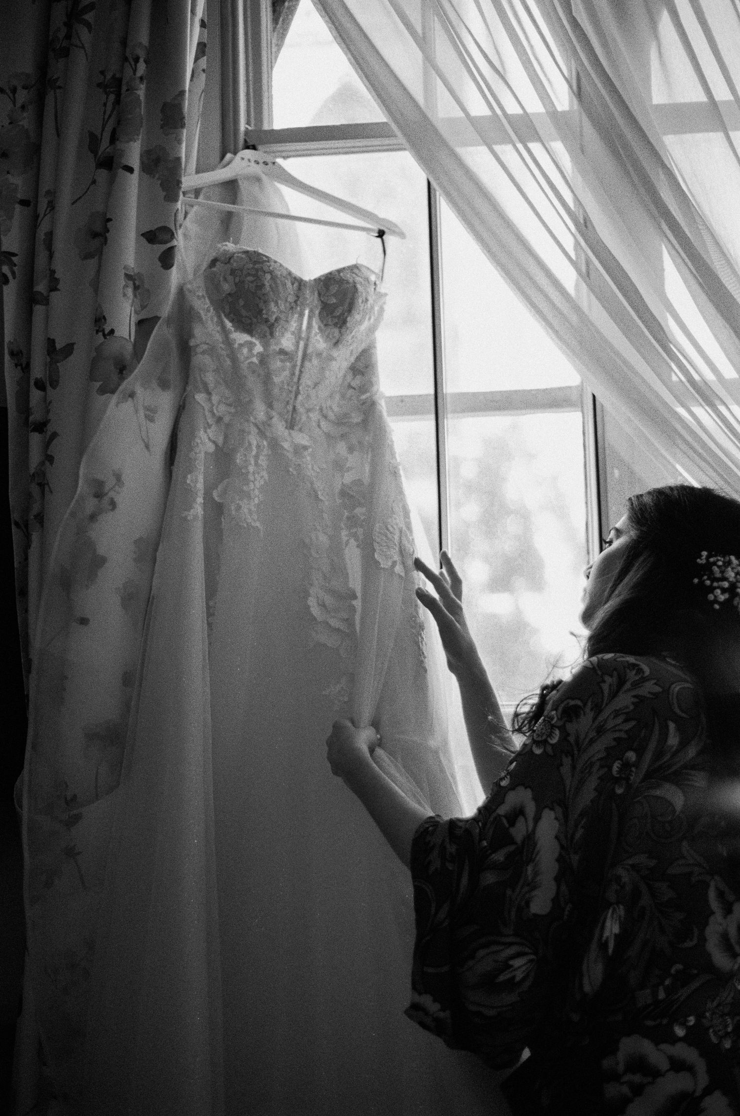 Black and white photo of bride admiring her lace wedding dress hanging by a window.