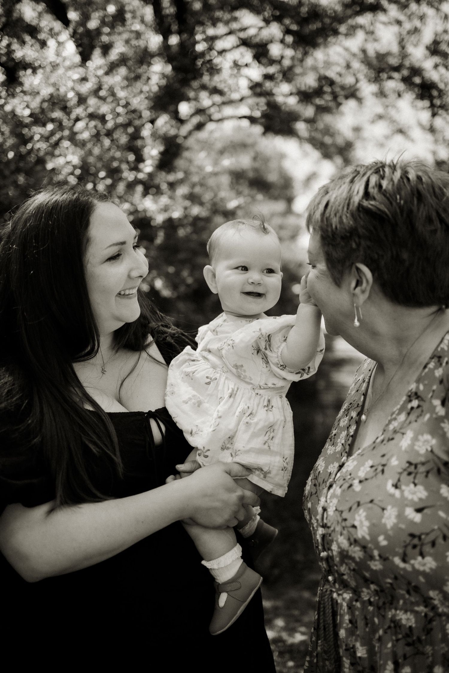 Black and white photo of mother holding happy baby reaching toward grandmother among garden trees.