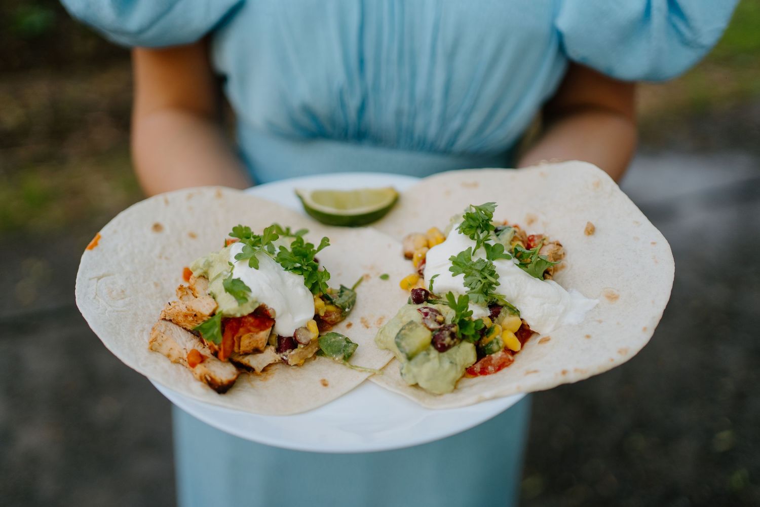 Person in blue dress holding a white plate with two loaded tacos garnished with lime.