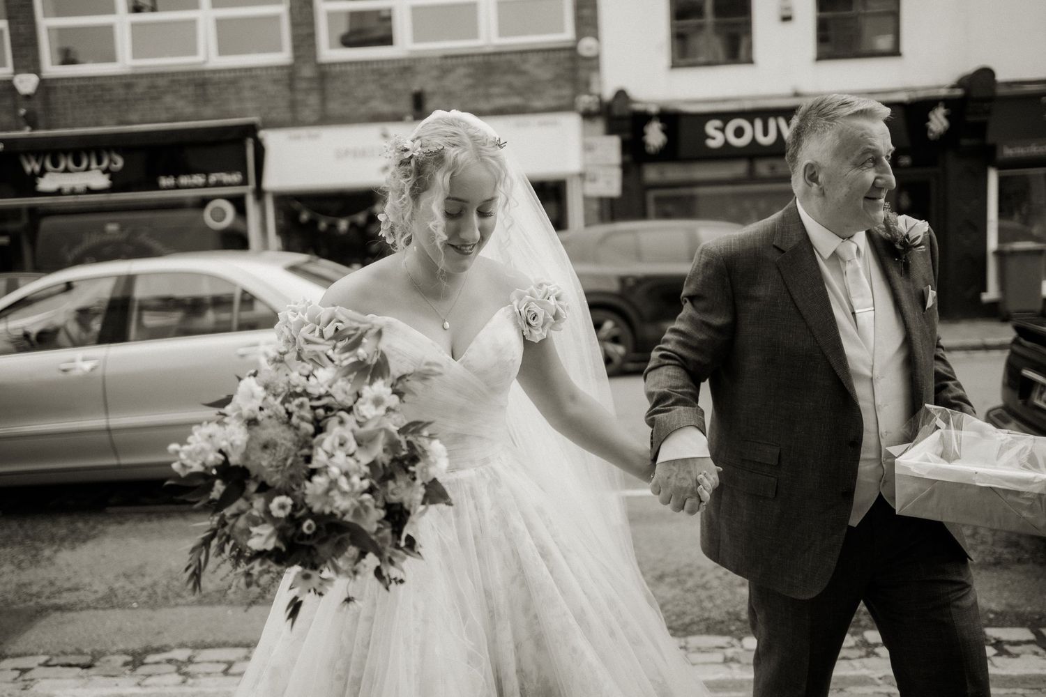 Black and white photo of bride walking with older gentleman holding bouquet on town street.