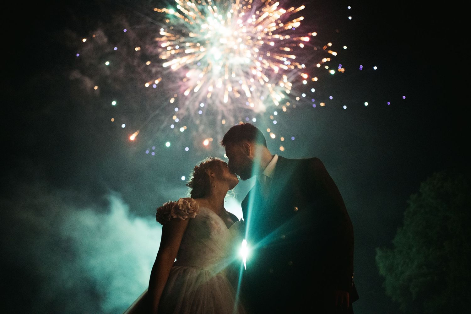 Wedding couple sharing a romantic kiss silhouetted against colorful fireworks in the night sky.