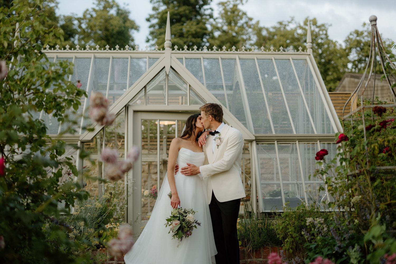 Bride and groom sharing romantic moment in front of glass greenhouse surrounded by roses.