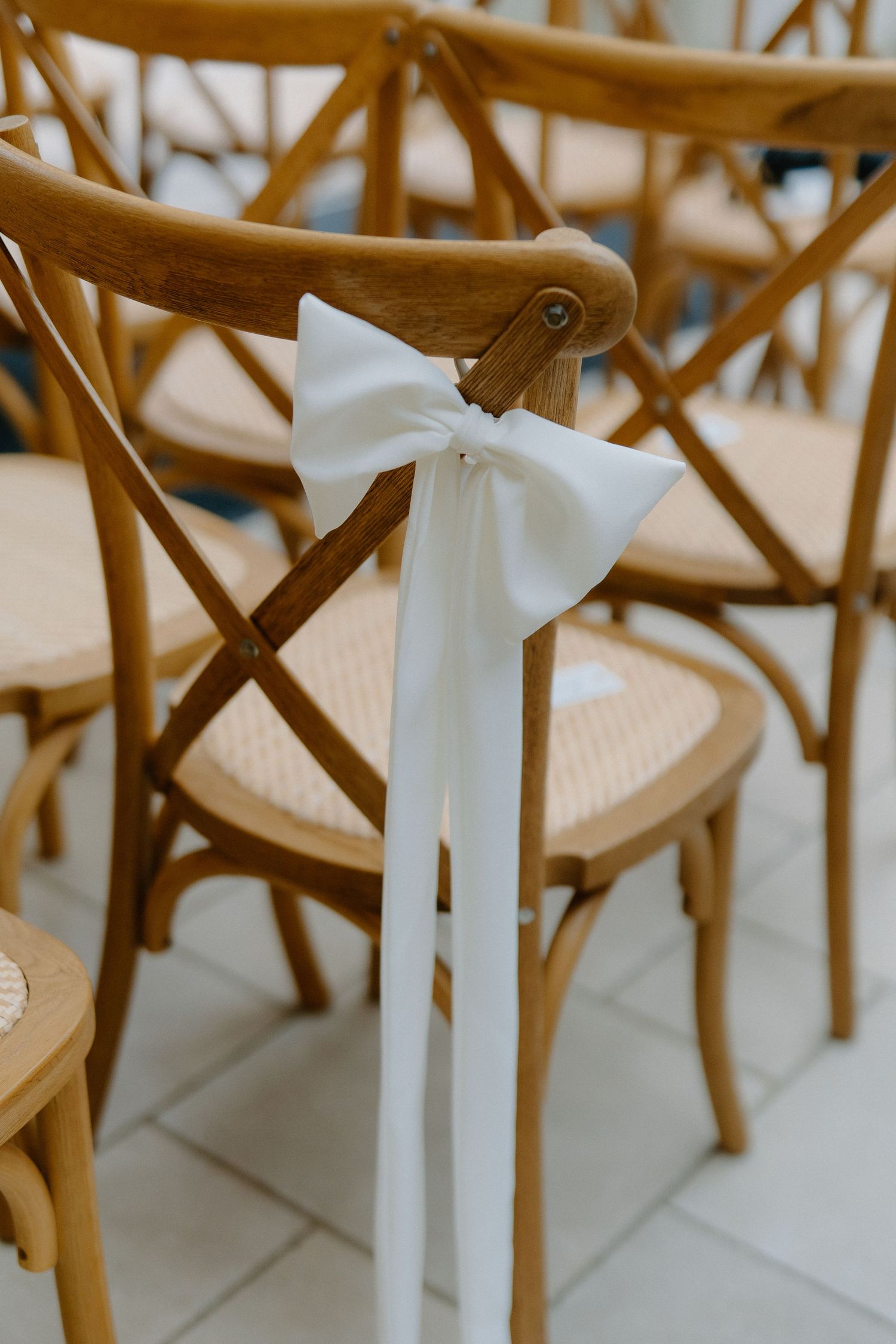 Wooden cross-back ceremony chairs decorated with white satin ribbon bows.