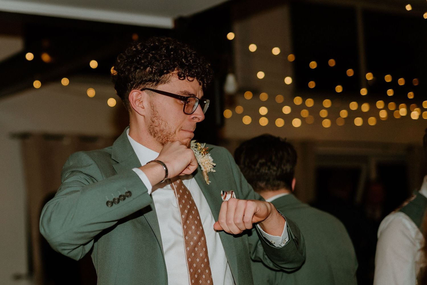 Stylish wedding guest in green suit and glasses dancing at a reception with fairy lights in background.