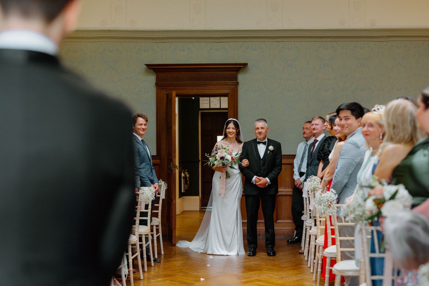 Bride in white gown walking down aisle with escort toward groom at wedding ceremony.