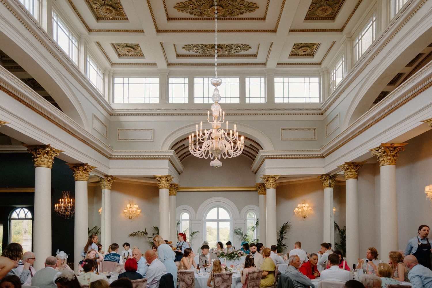 Elegant wedding reception hall with high ceilings, crystal chandelier, gold columns, and seated guests.