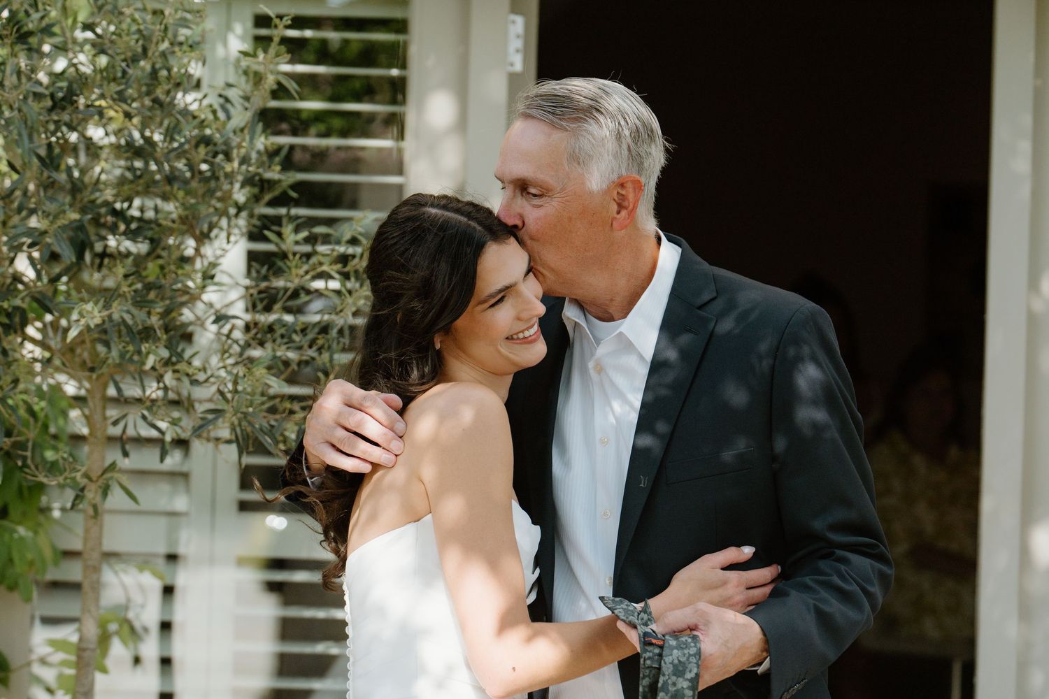 Father of the bride kisses her cheek lovingly as she smiles in her wedding dress.