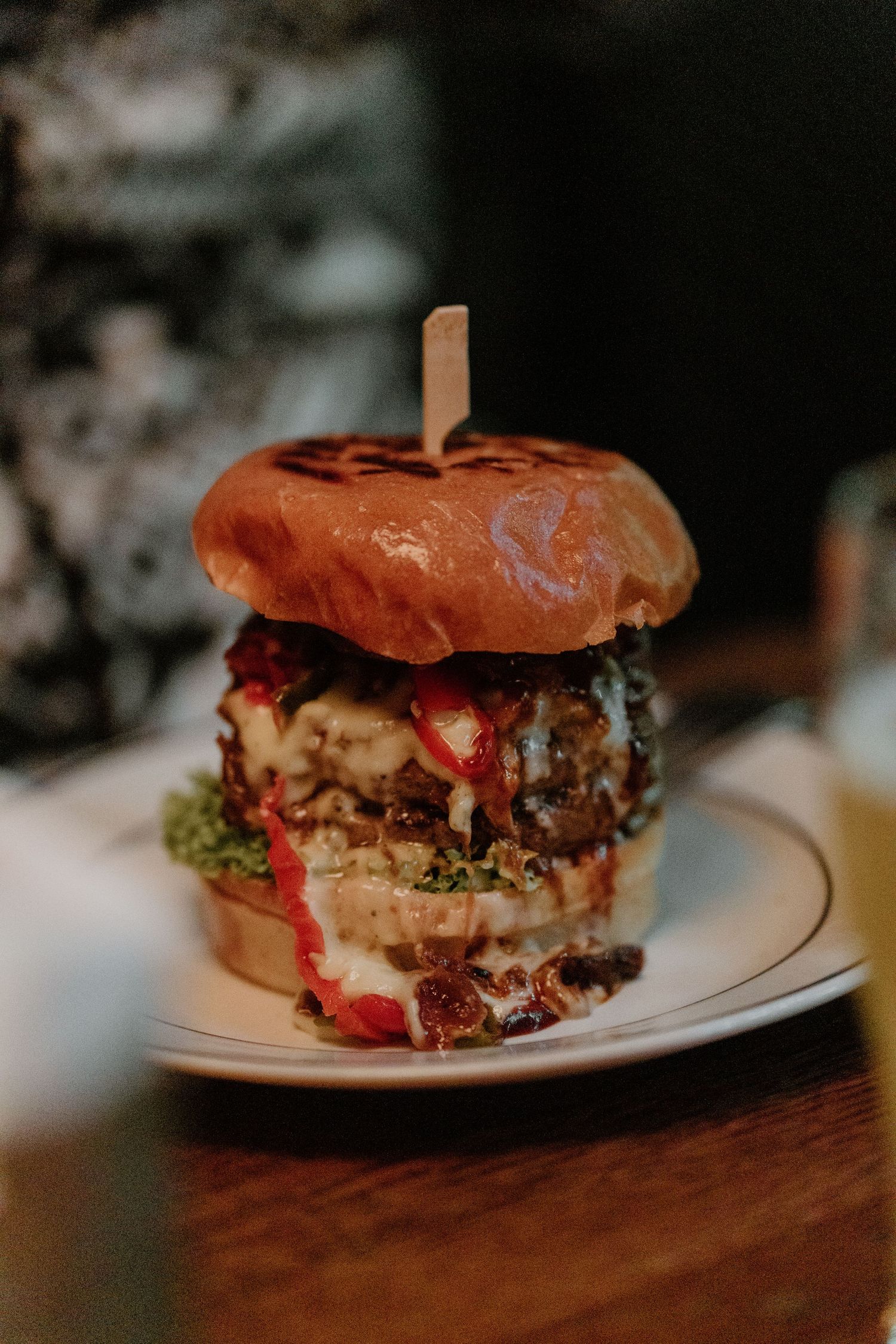 A juicy gourmet burger stacked high with melted cheese, red sauce, and toppings on a brioche bun served on a white plate.