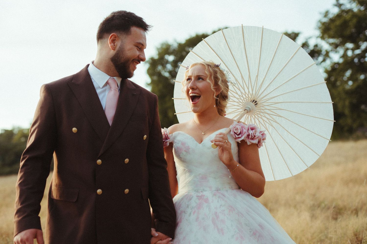 A couple in wedding attire walks through a sunny field, with the bride holding a white decorative parasol.