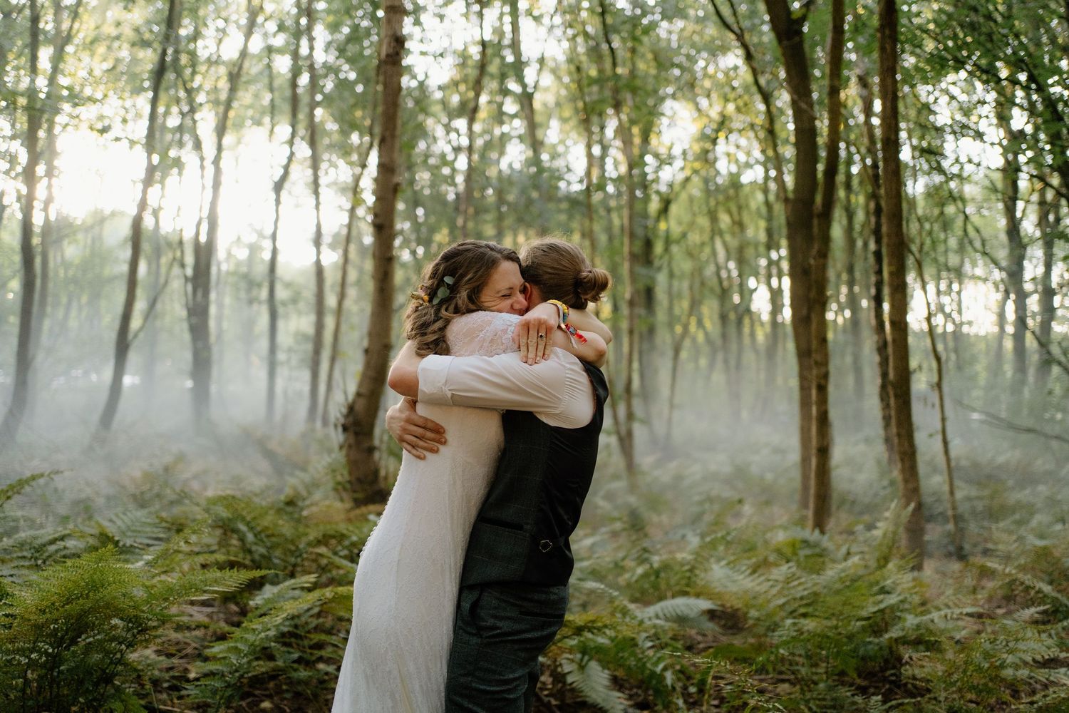 Newlyweds share a tender embrace in a misty forest surrounded by lush green ferns and tall trees.