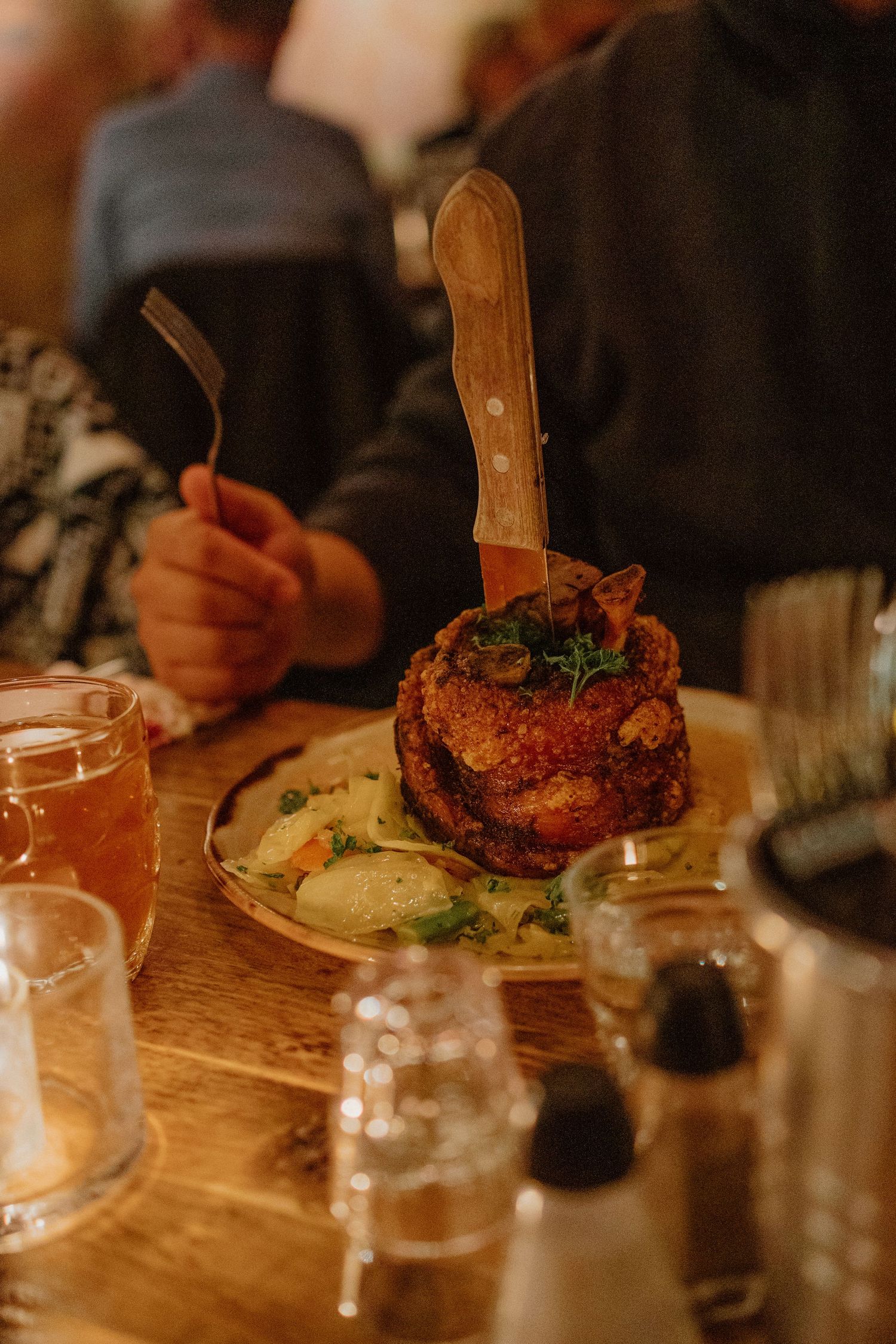 Close-up of a gourmet pork knuckle stack with garnish on a wooden table in warm lighting.