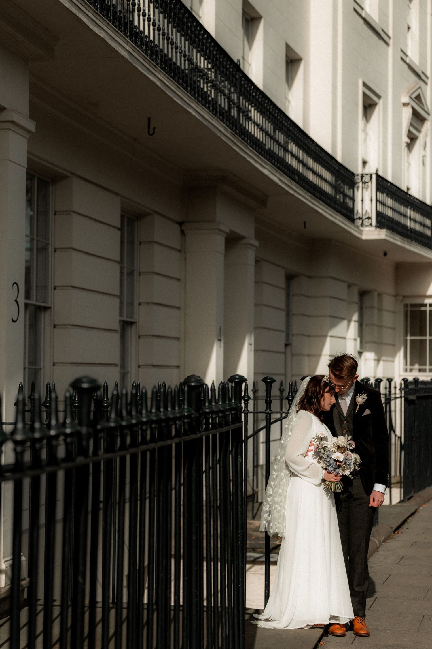 A couple in wedding attire stand intimately together against a white Georgian townhouse with ornate iron railings in London.