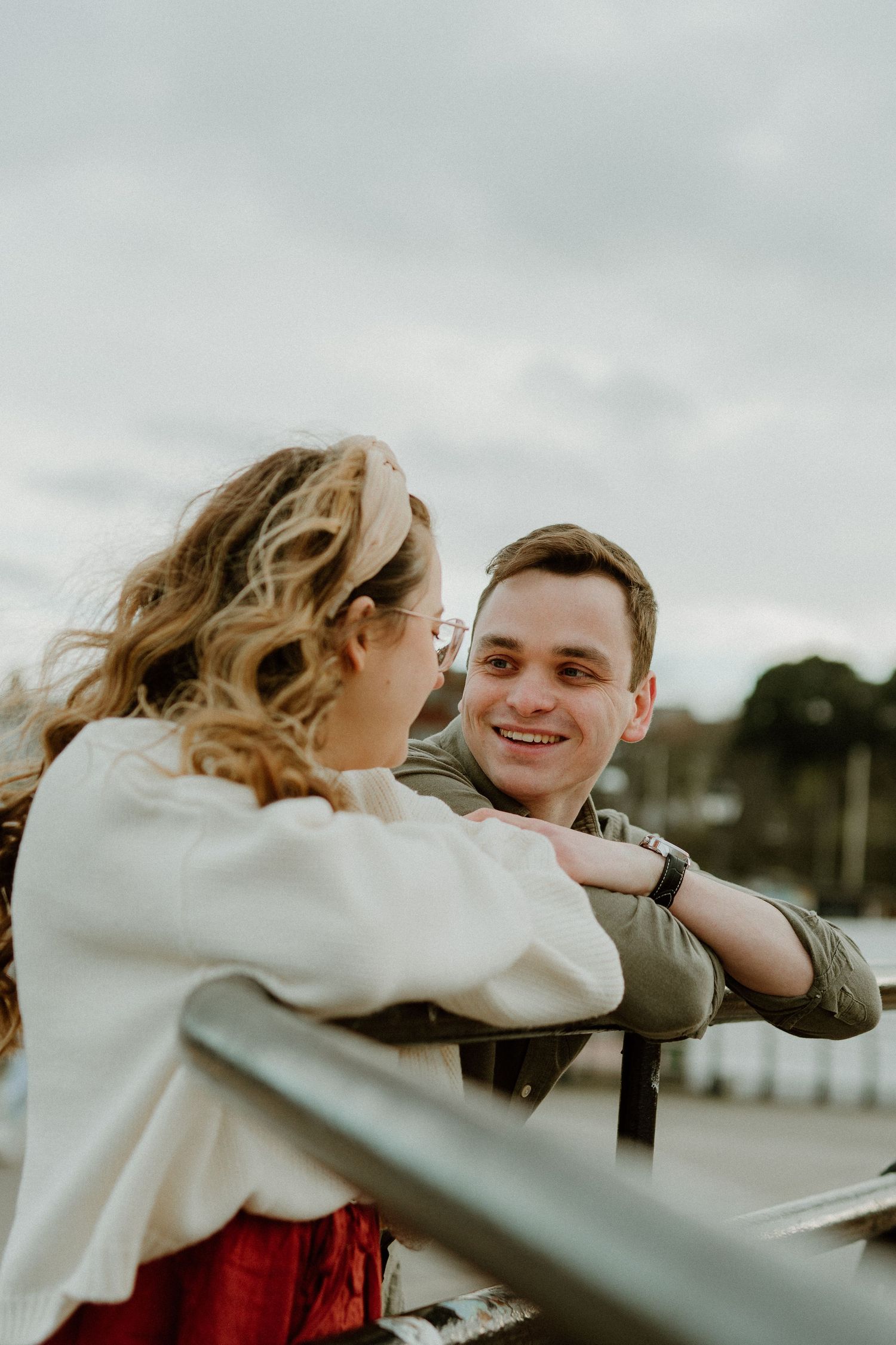 A couple shares a tender moment together while leaning against a railing during sunset with moody overcast skies.
