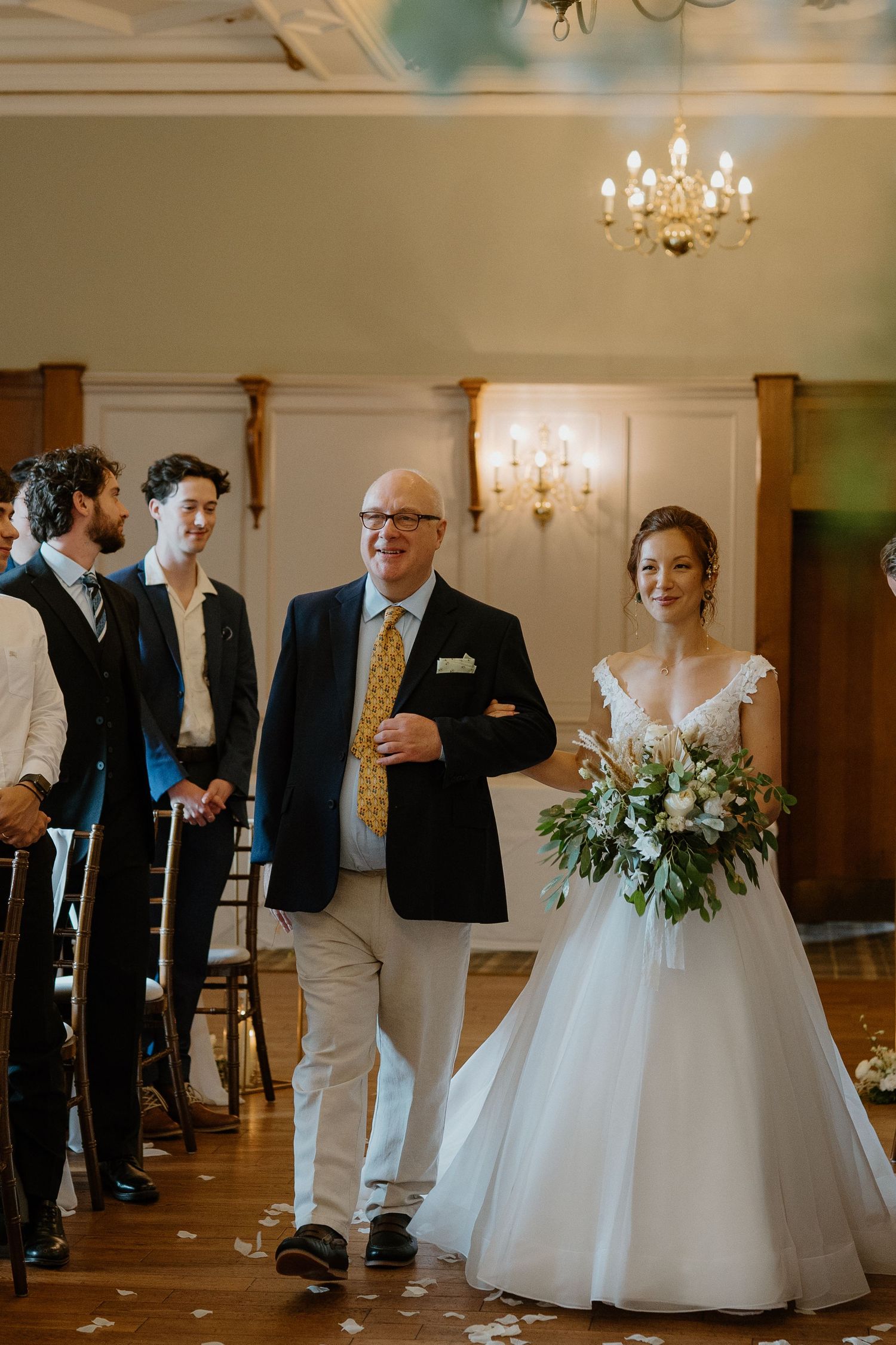 Smiling bride walks down aisle with father in elegant chandelier-lit wedding ceremony hall.