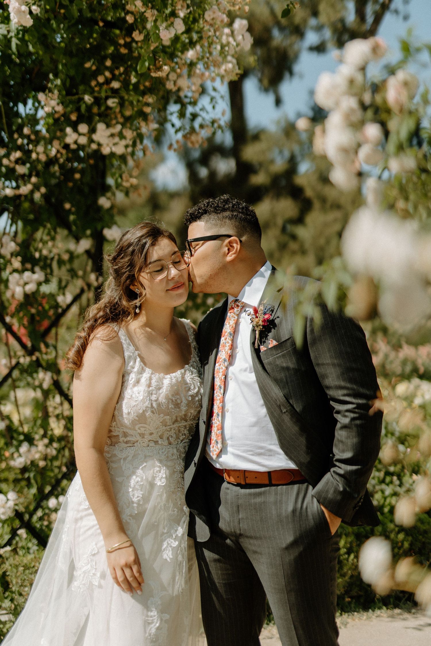 Groom kissing bride on the cheek beneath a canopy of blooming white roses in an outdoor garden.