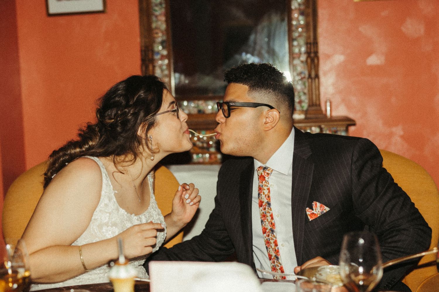 Newlyweds share a kiss while seated at dinner table, groom wearing floral tie and navy suit.