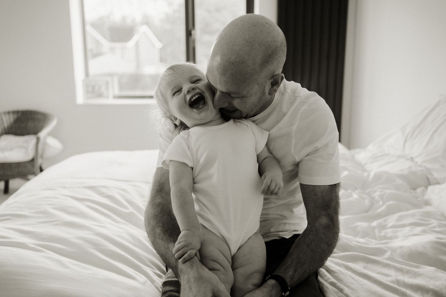 Black and white photo of a bald father laughing with his joyful baby on a bed.