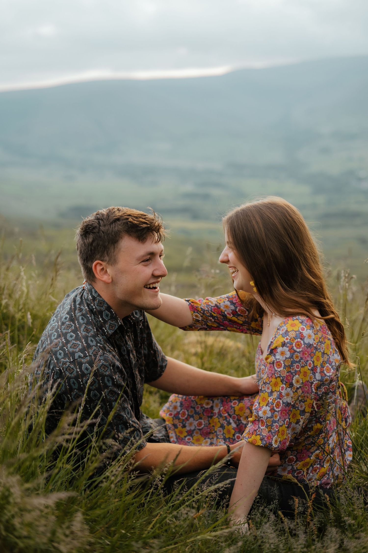 Happy couple laughing and sitting together in tall grass with misty mountain backdrop.