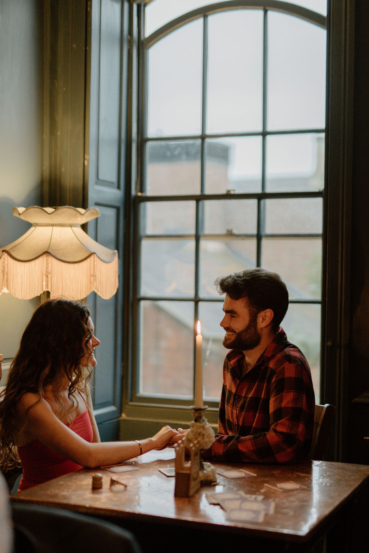 Couple holding hands across a candlelit table by a large window in a cozy vintage restaurant.