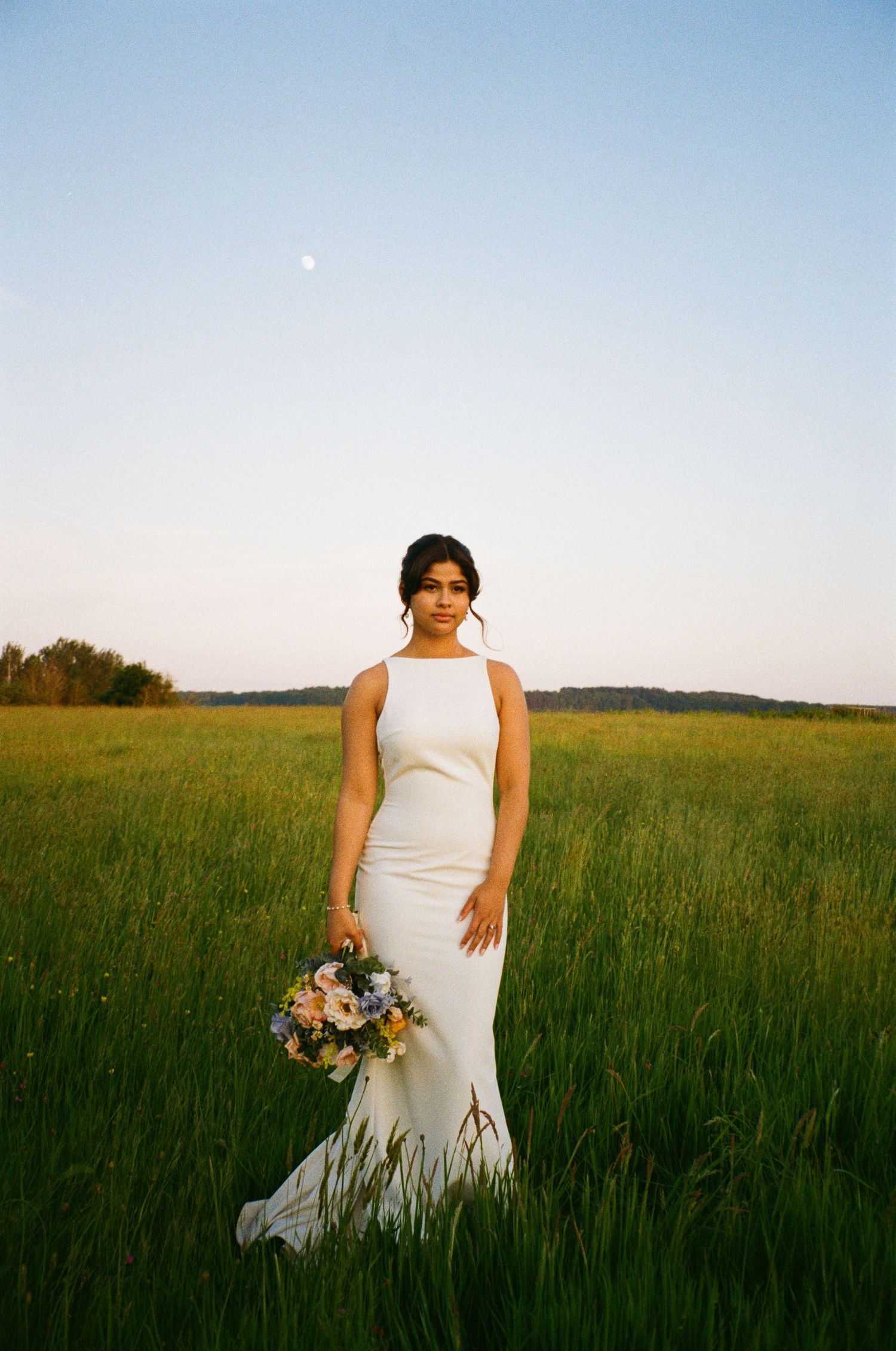 Elegant bride in sleek white gown holds pastel bouquet while standing in vast green field under moonlit sky.