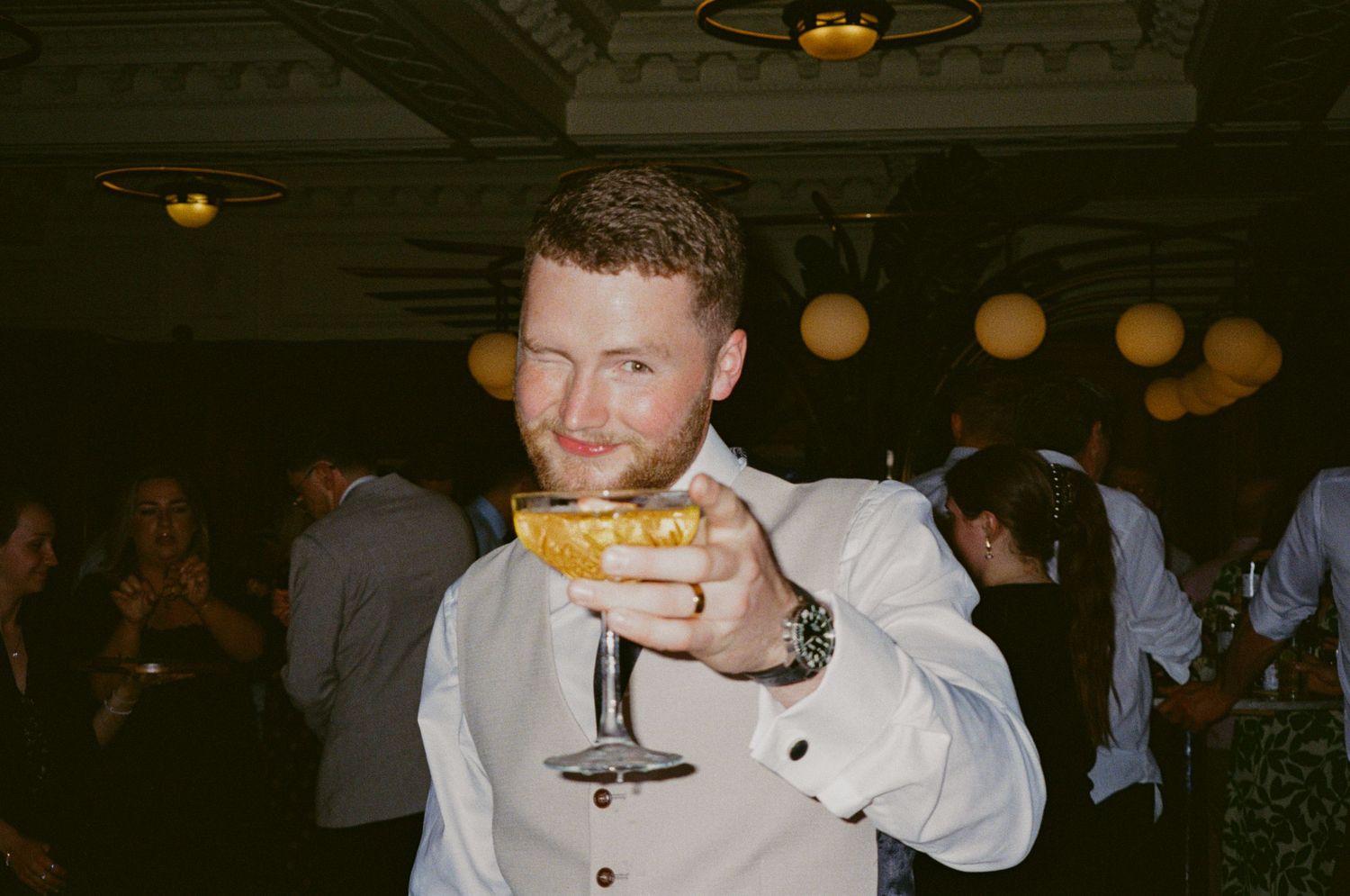 Smiling red-haired man holding a cocktail glass at a lively wedding reception.