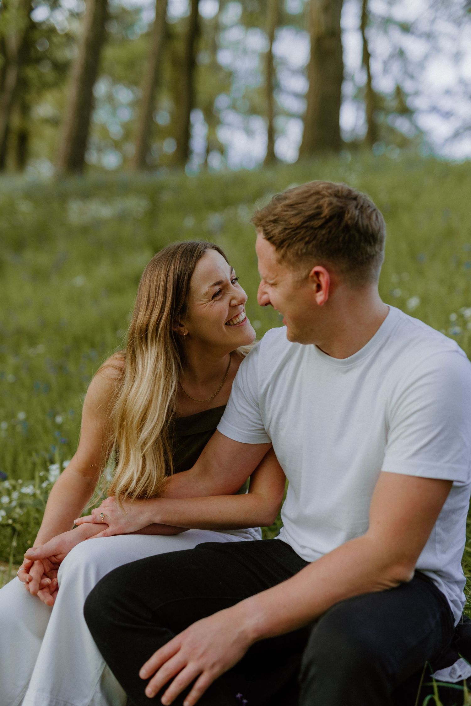 Smiling couple sitting close together in a blooming green forest meadow, looking at each other.