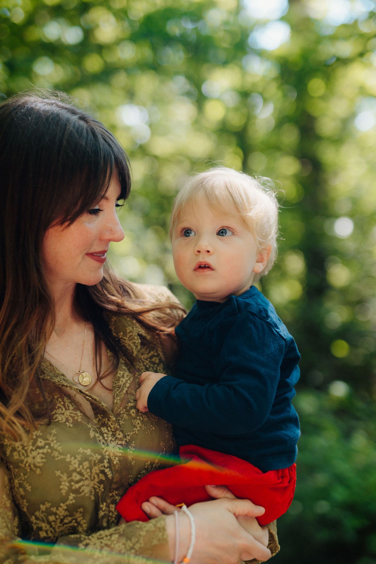 A dark-haired mother holds a wide-eyed blonde toddler in a sunlit green forest.