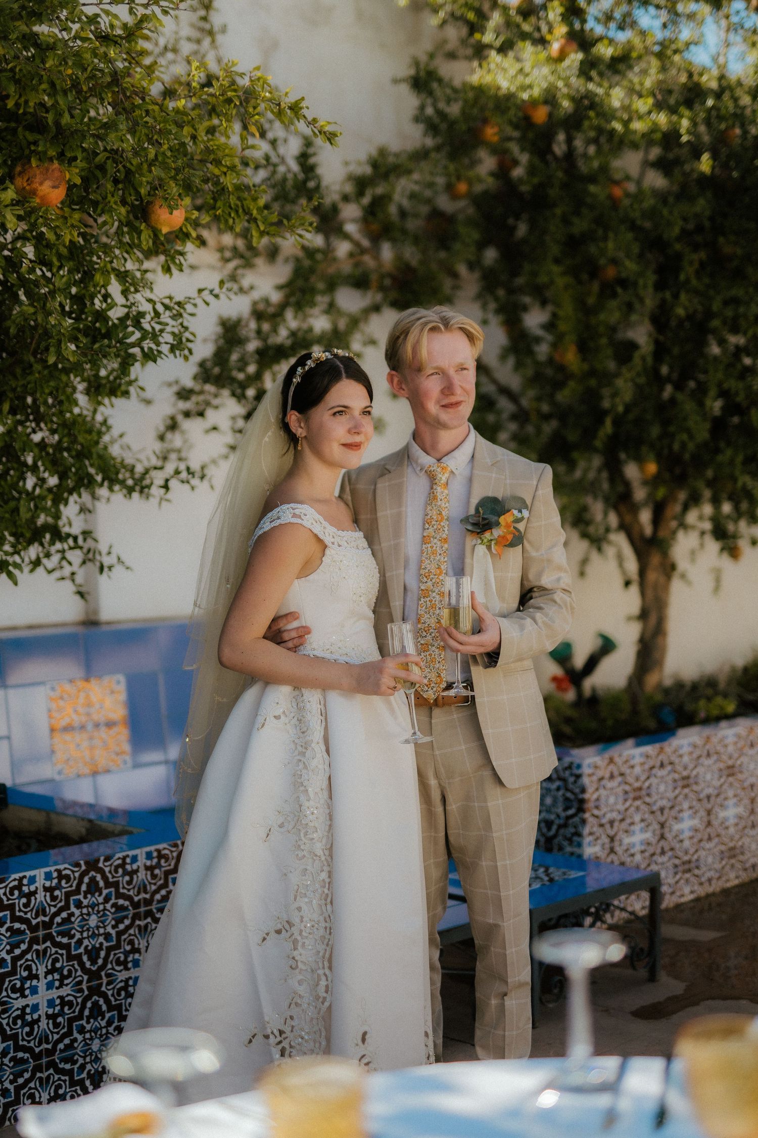 Bride and groom pose together under orange tree holding champagne glasses at reception.