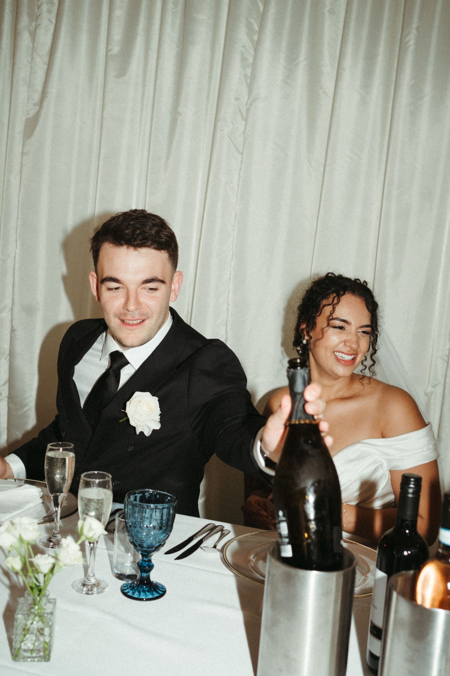Bride and groom smile and laugh together at their wedding reception table with champagne and flowers.