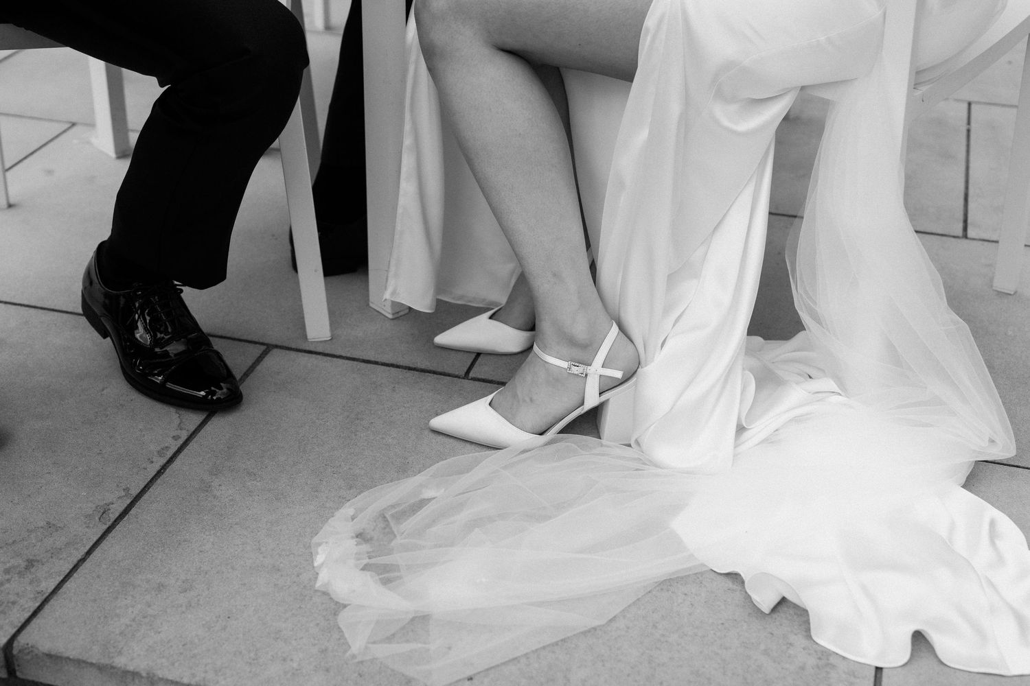 Black and white photo of bride in white heels and groom in black dress shoes seated at wedding ceremony.
