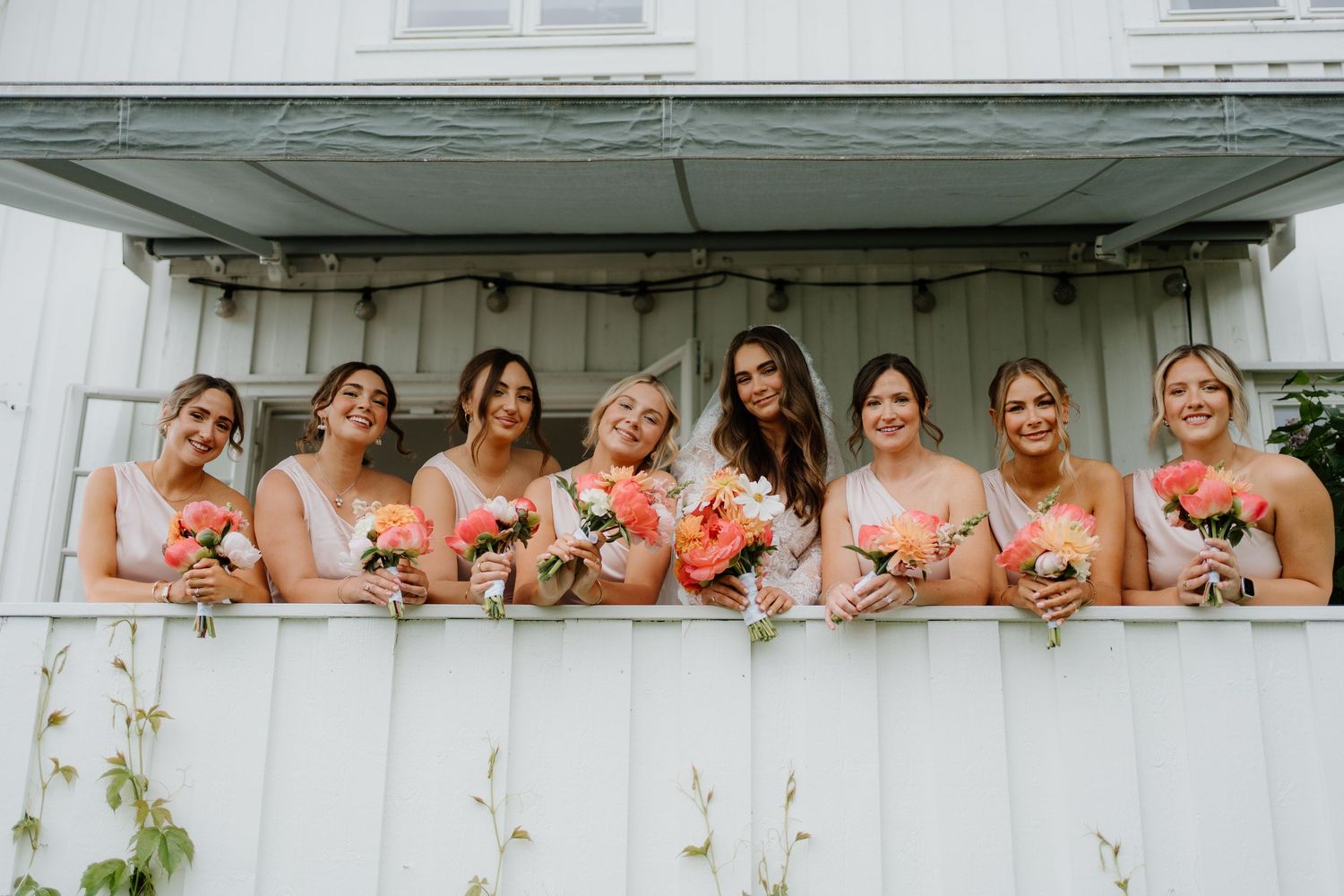 Seven bridesmaids in blush dresses holding coral peony bouquets smiling on a porch.