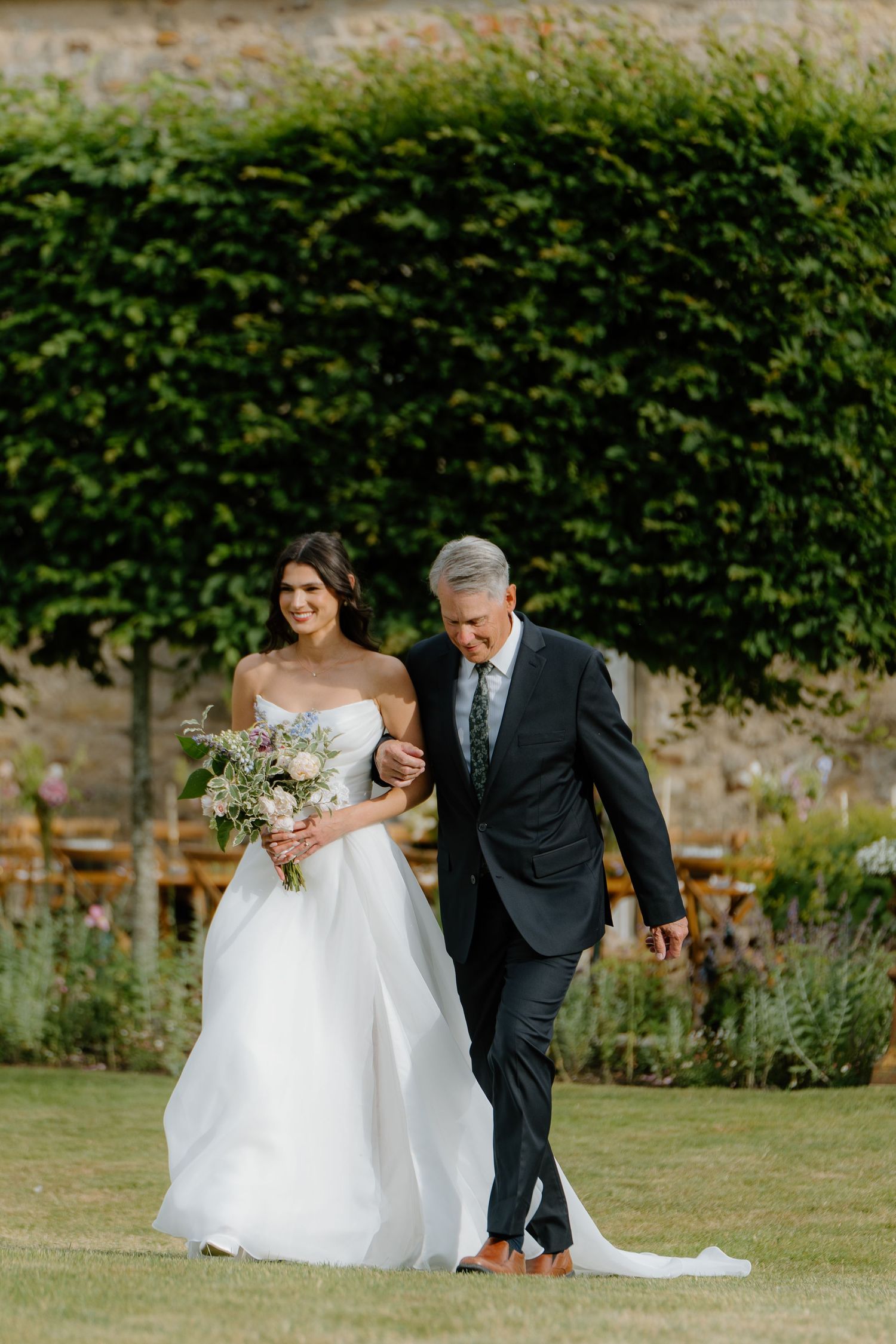 Bride in white ballgown holding pastel bouquet walks down aisle with father at outdoor wedding.
