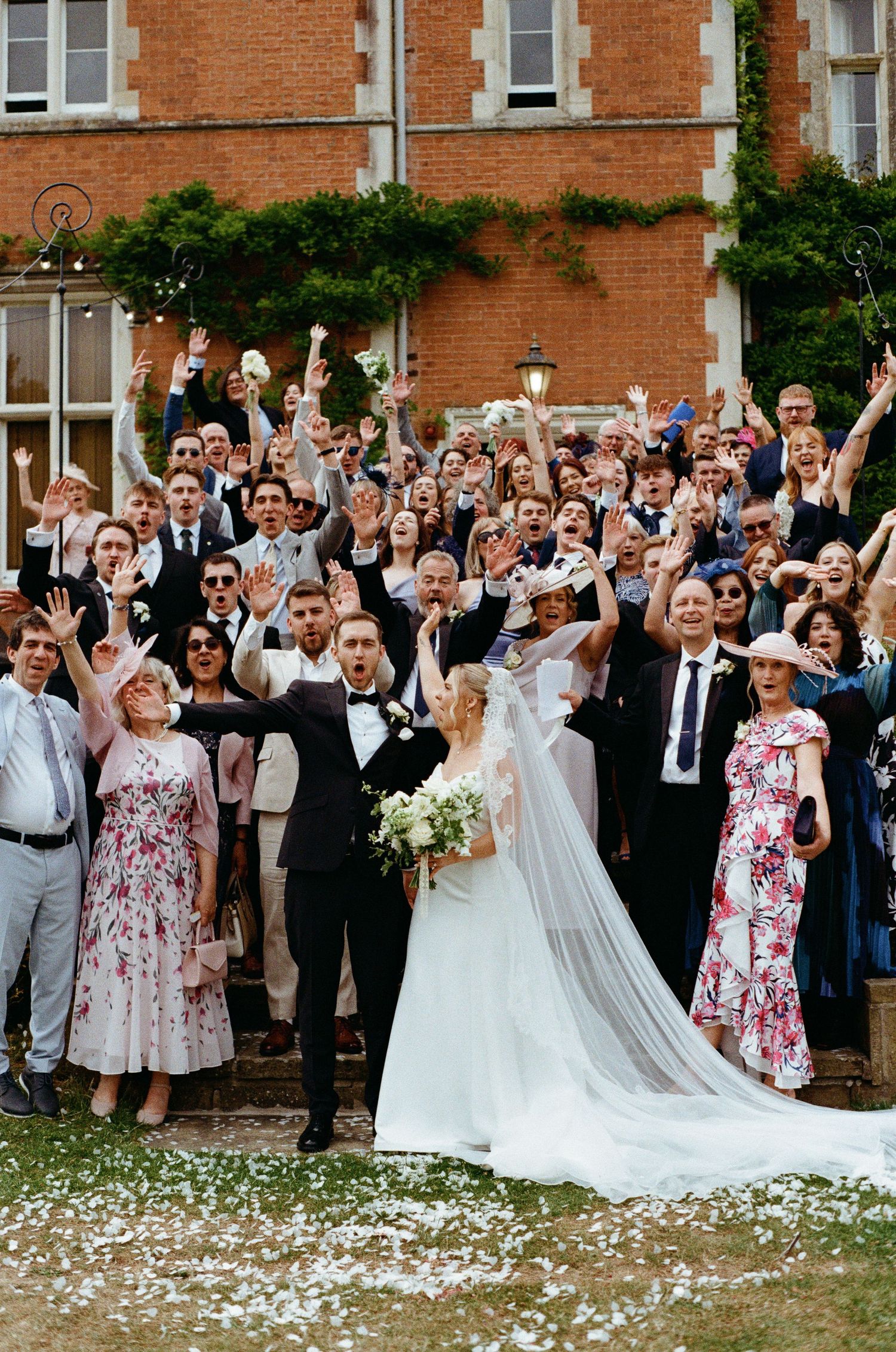 Joyful wedding couple surrounded by cheering guests throwing confetti outside historic brick manor.
