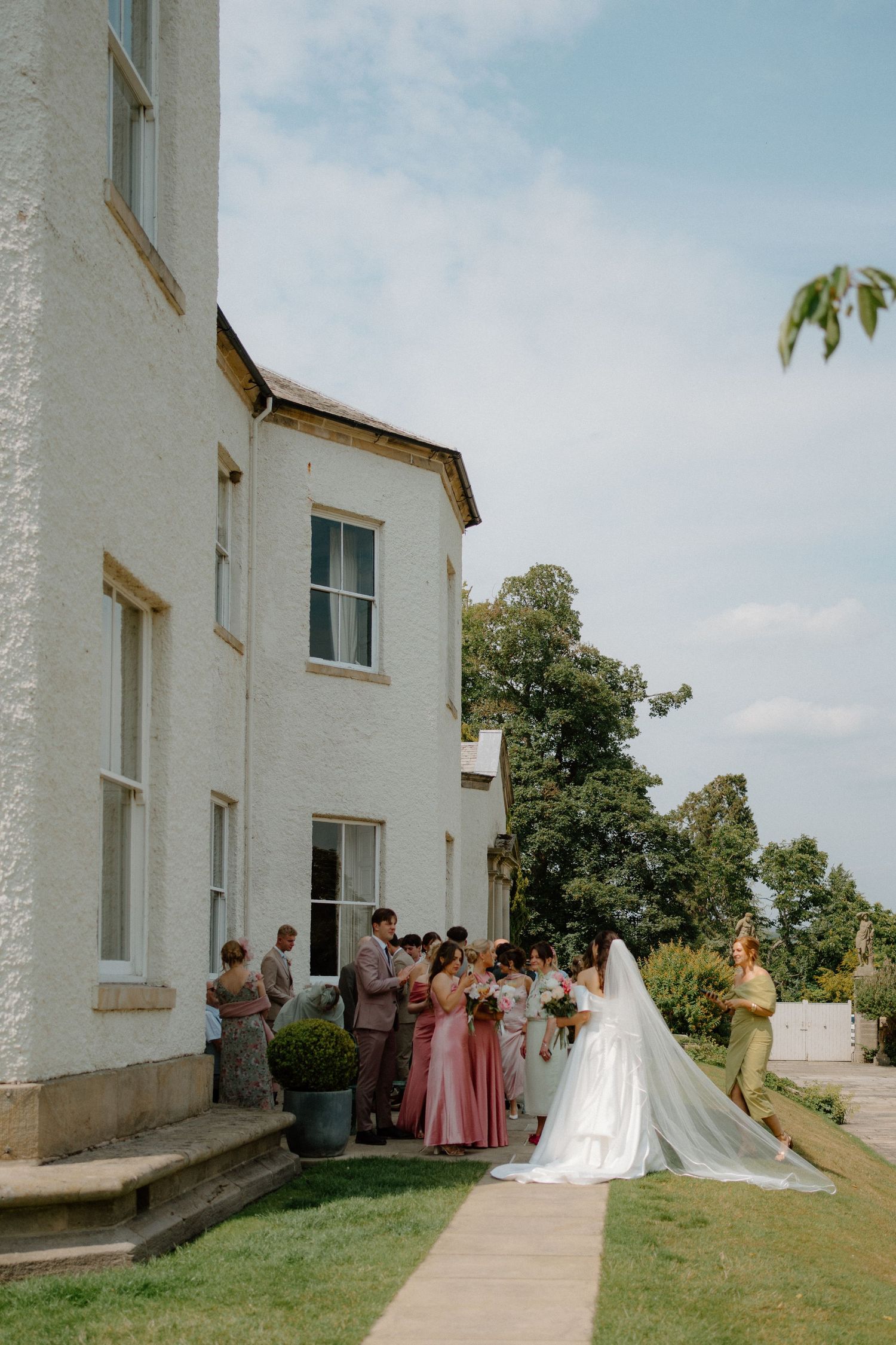 Bride in flowing white gown with long veil walking toward wedding party outside white manor.