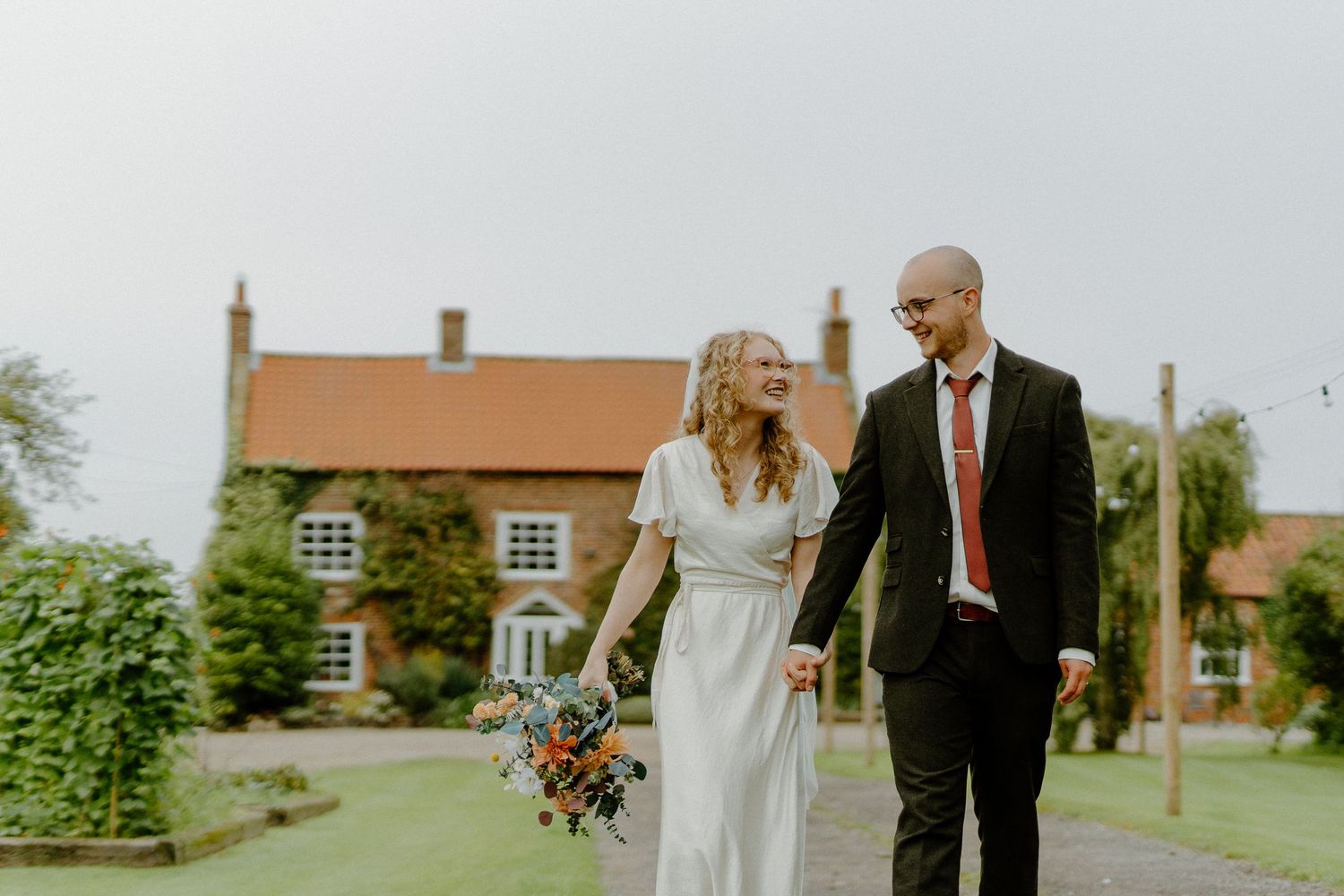 Bride and groom walking hand in hand outside a rustic farmhouse venue, bride holding a wildflower bouquet.