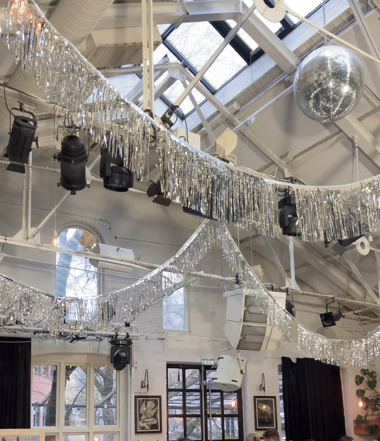 Silver disco ball and sparkling tinsel decorations hang from an industrial ceiling with stage lighting fixtures.