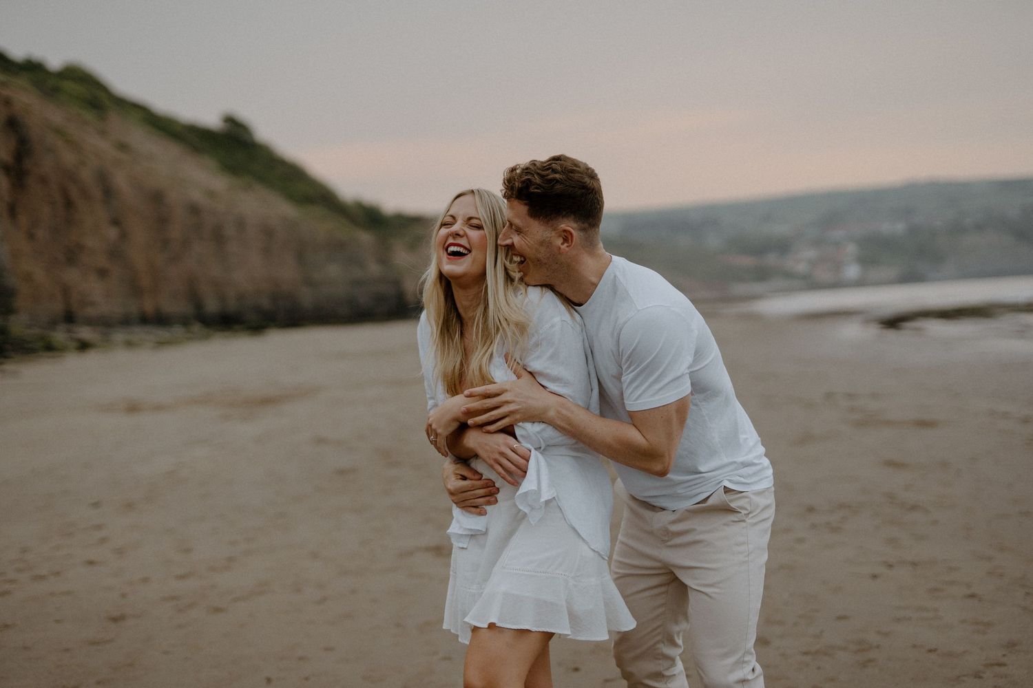 A happy couple laughs together while embracing on a secluded beach at sunset with cliffs in the background.