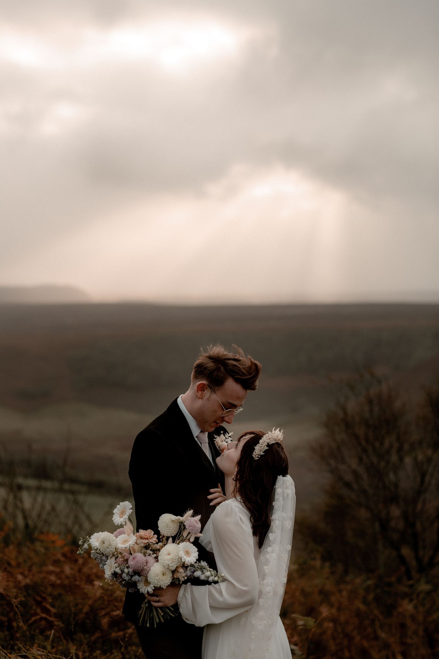 Atmospheric wedding portrait with dramatic cloudy sky and rolling hills in background.