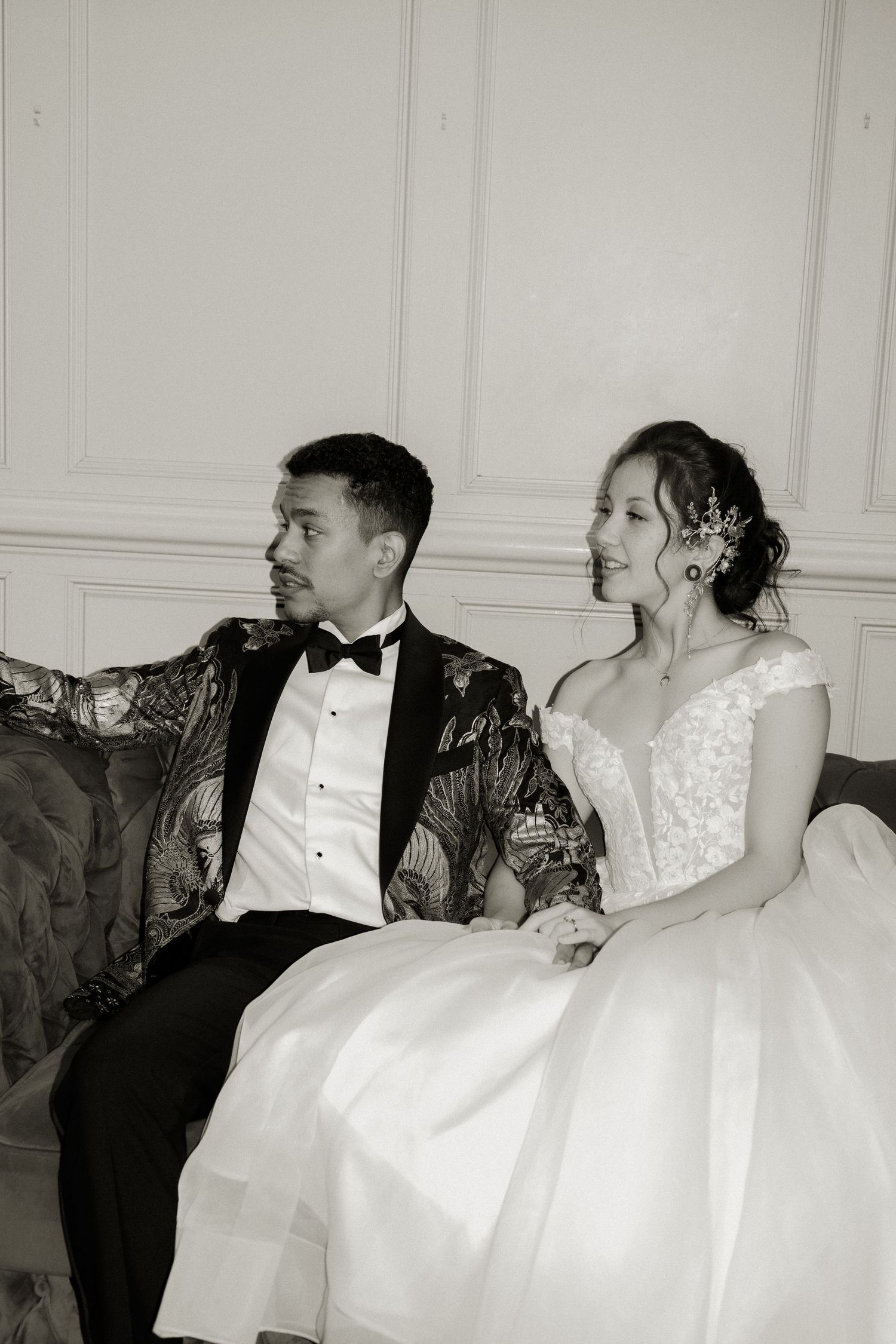 Black and white photo of bride and groom sitting together on ornate sofa in elegant wedding attire.