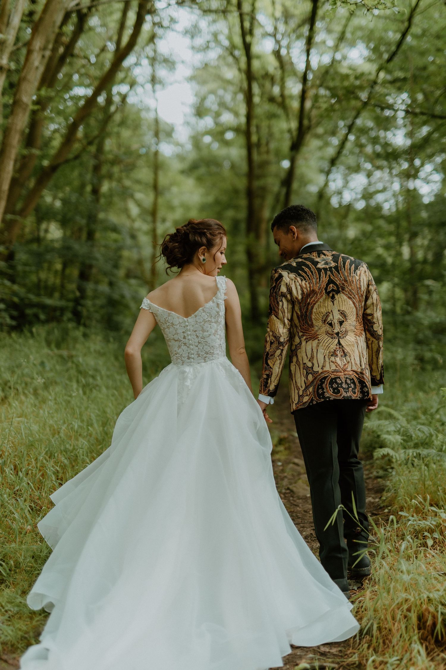 Bride in elegant white ballgown and groom in gold batik jacket walk hand-in-hand through forest.